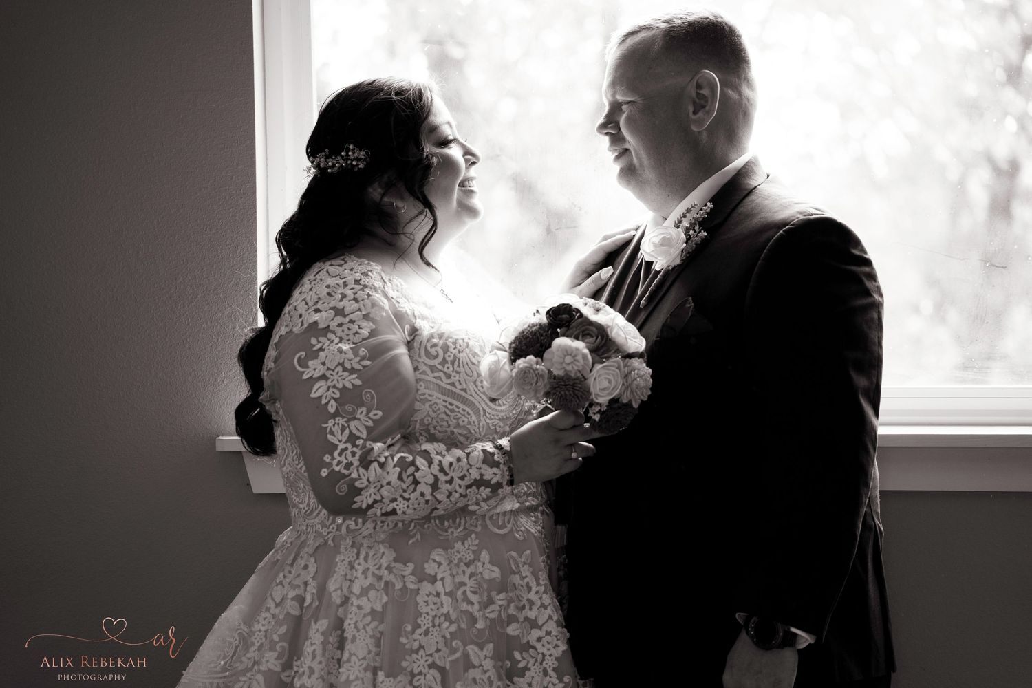 A romantic black and white moment between newlyweds by a bright window.