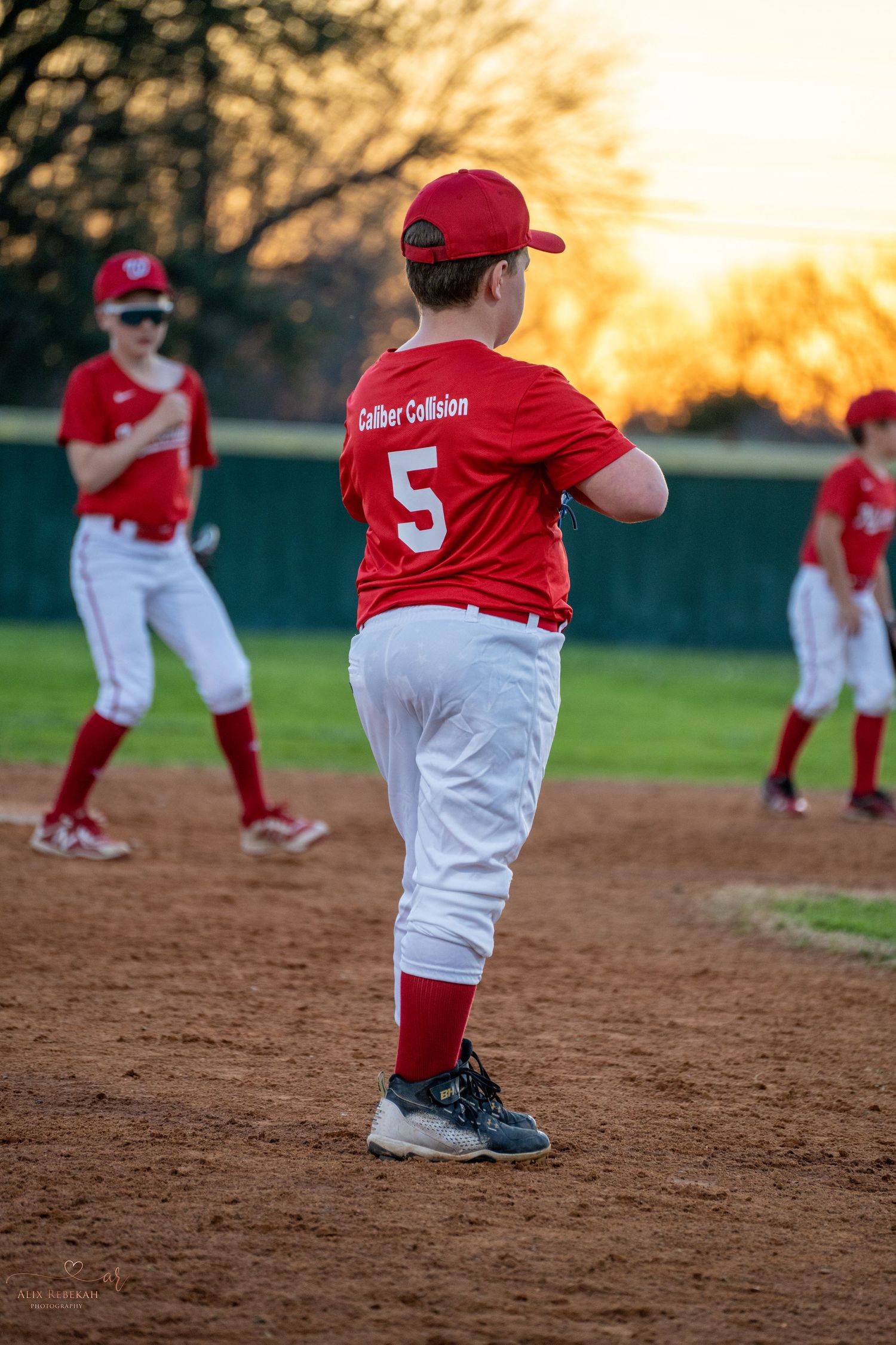 Youth baseball player in red uniform #5 stands on dirt infield during sunset at baseball game practice session.