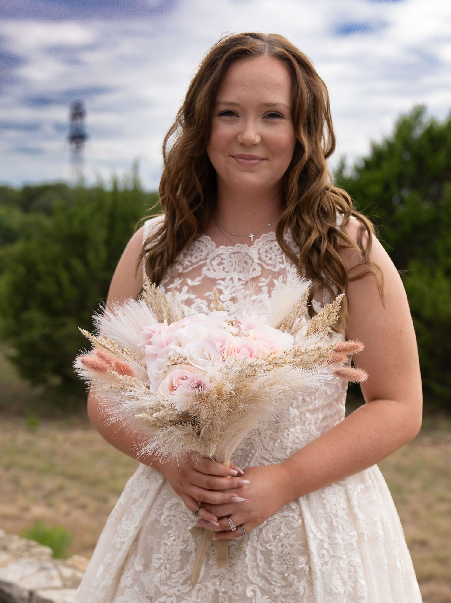 Bride in lace wedding dress holding pink floral bouquet with pampas grass outdoors.