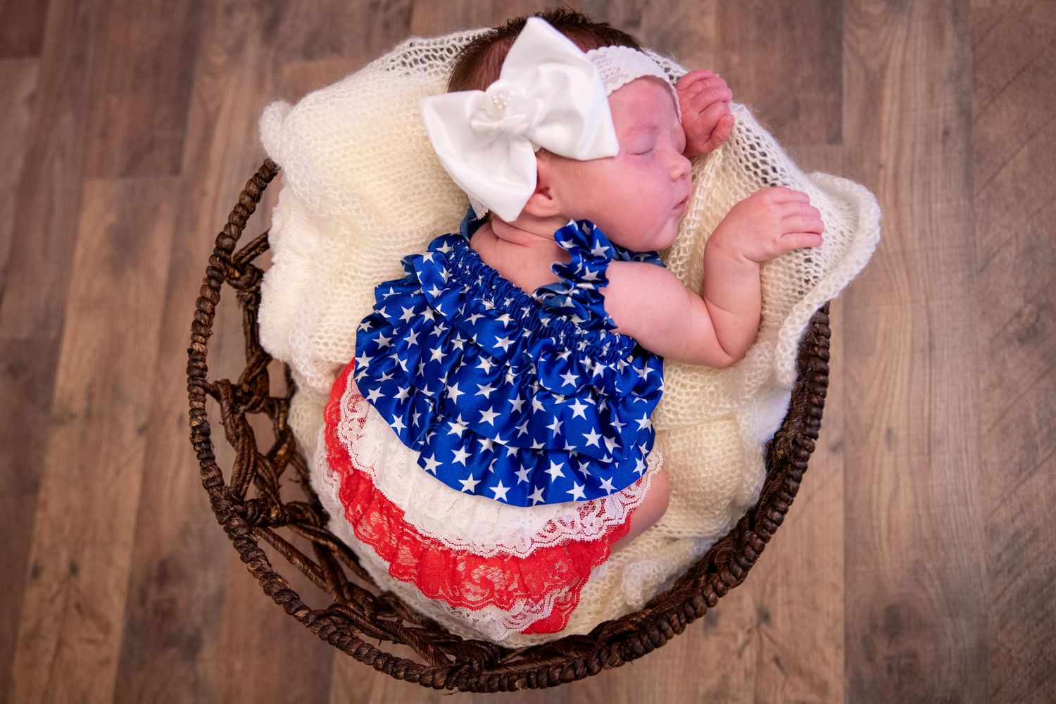 Newborn baby in patriotic outfit sleeping in wooden basket on hardwood floor.