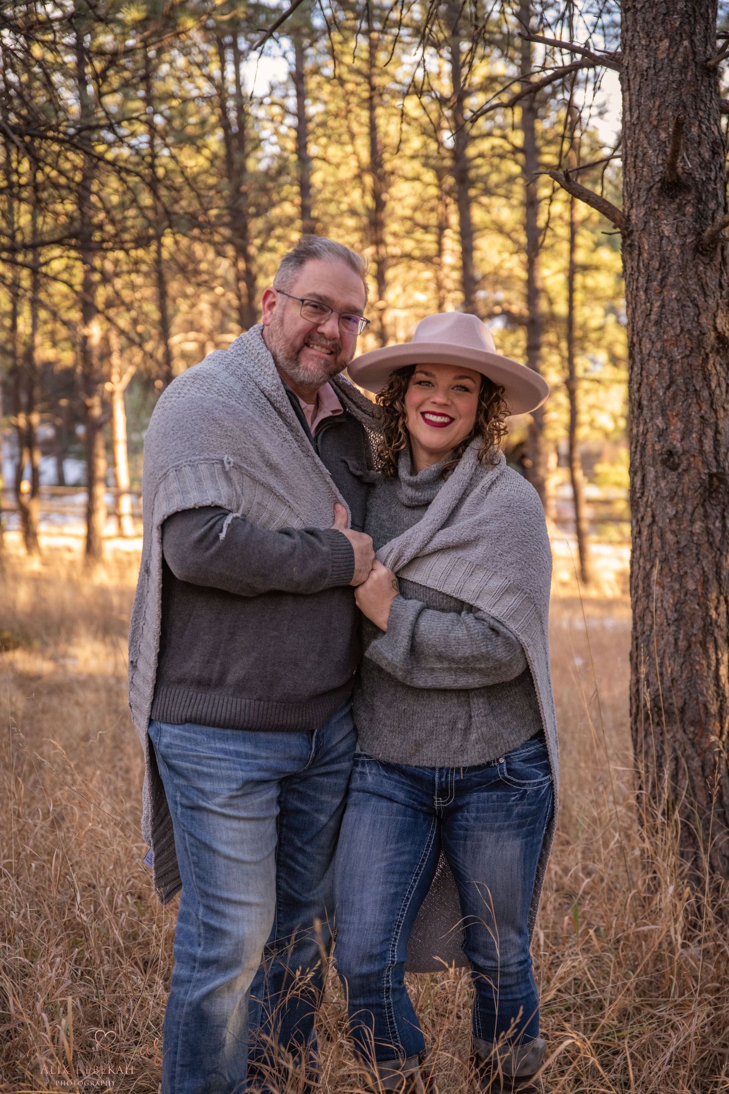 Happy couple in gray sweaters laughing together against winter forest backdrop.