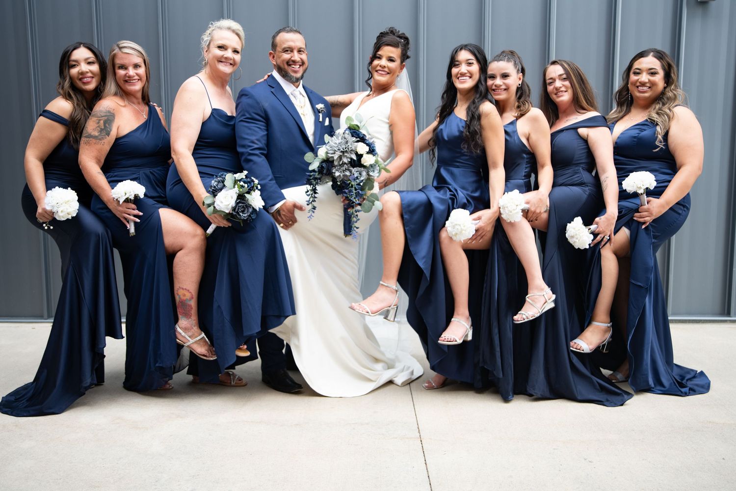 A wedding party poses in coordinated navy blue dresses and grey suits outdoors.
