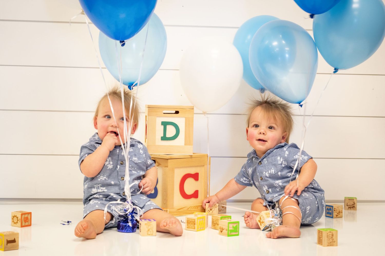 Twin babies in matching outfits sitting by alphabet blocks and blue balloons.