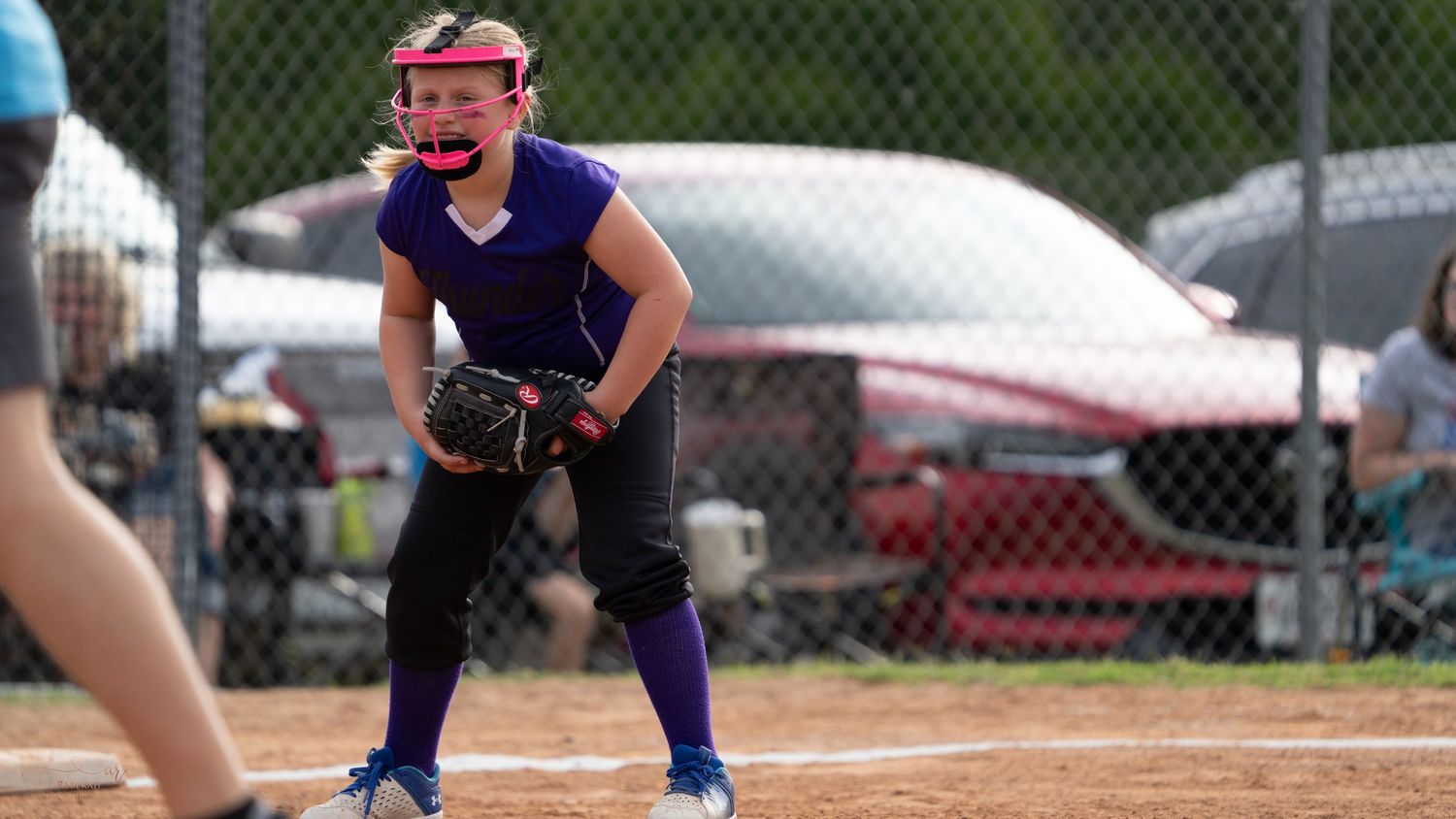 Softball player in blue jersey and face mask crouches in ready position with mitt at first base during game.