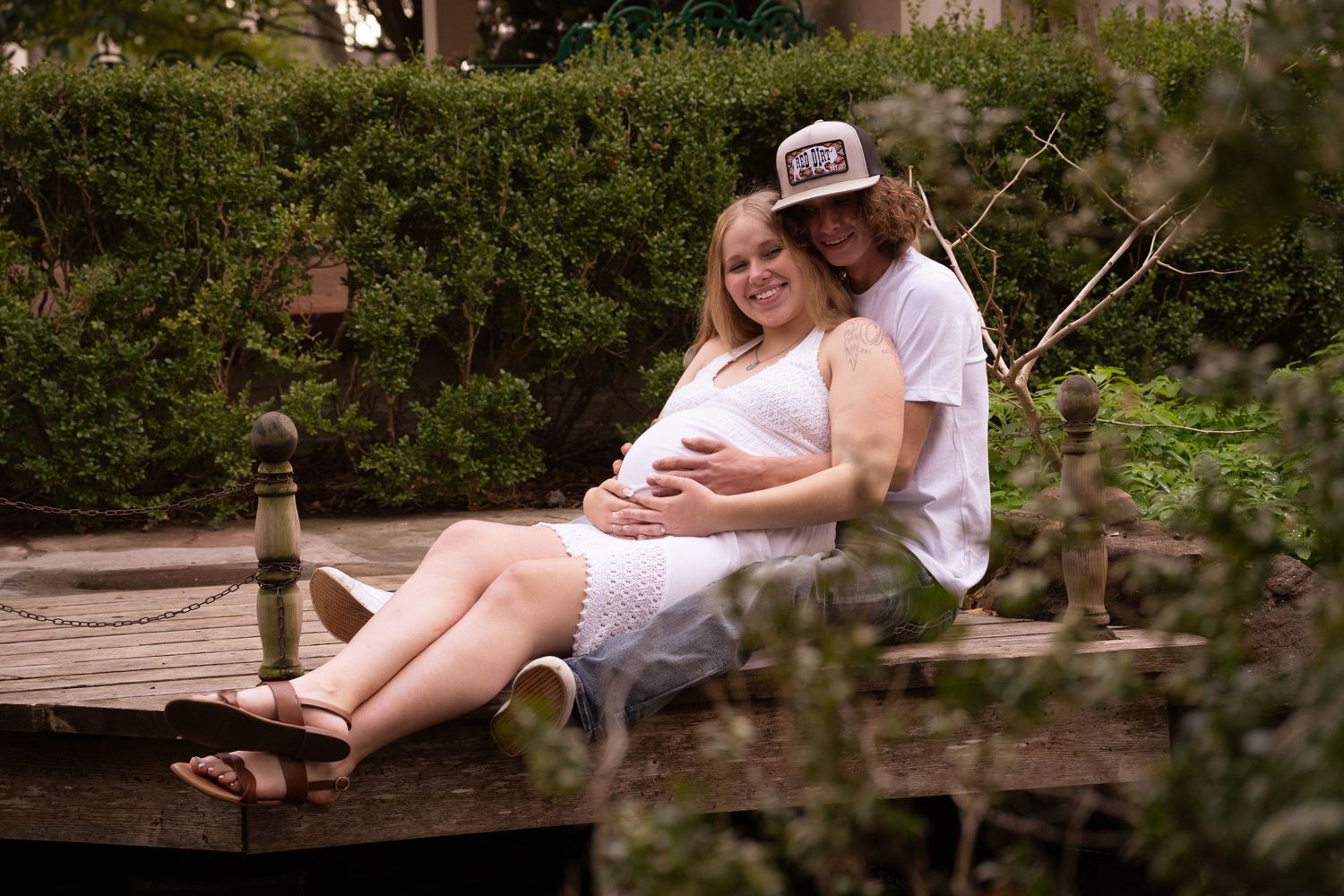 A couple embraces on a wooden dock surrounded by lush greenery during a maternity photo session.