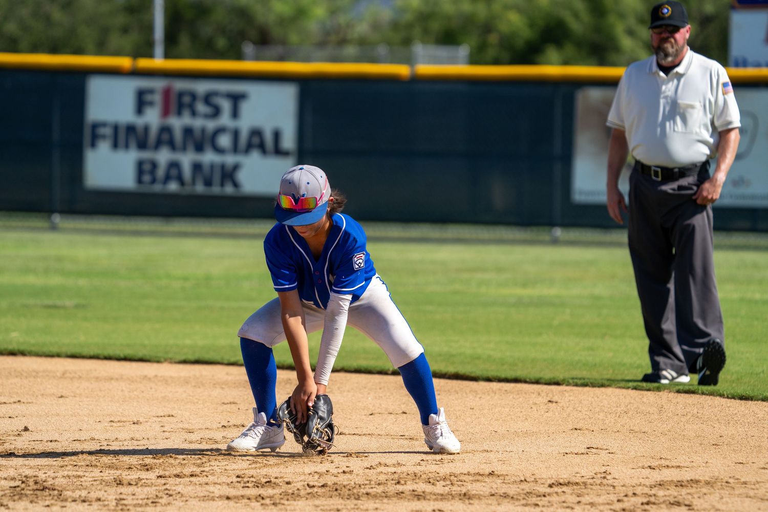 Baseball infielder in blue uniform crouches in ready position while umpire watches from behind at First Financial Bank field.