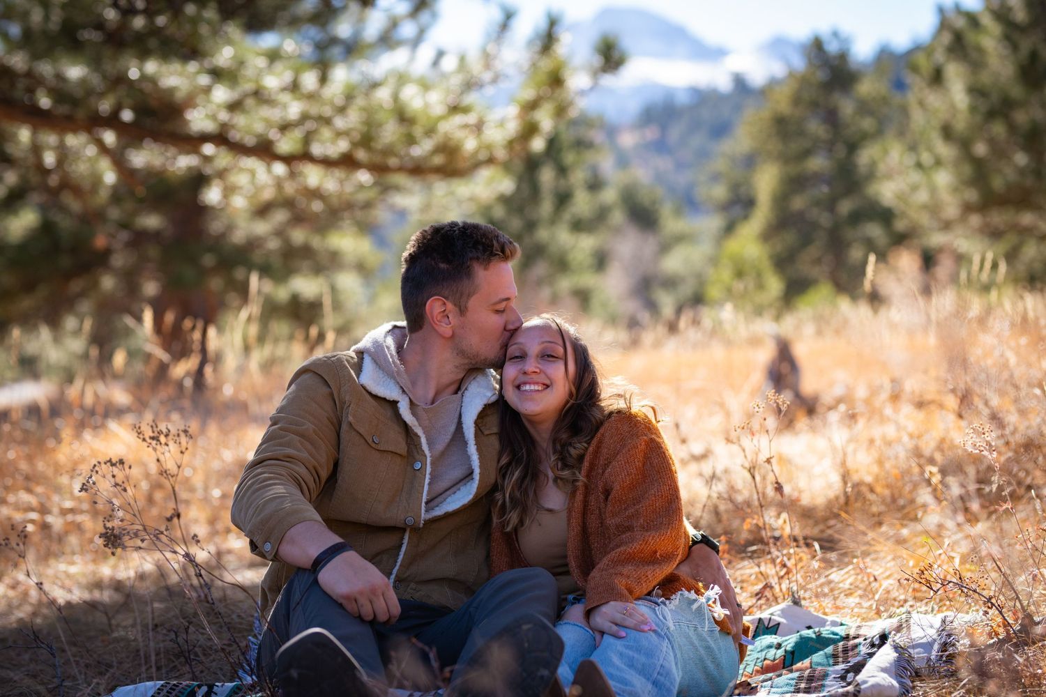 Two people share a joyful moment while sitting together outdoors against a backdrop of pine trees and mountains.