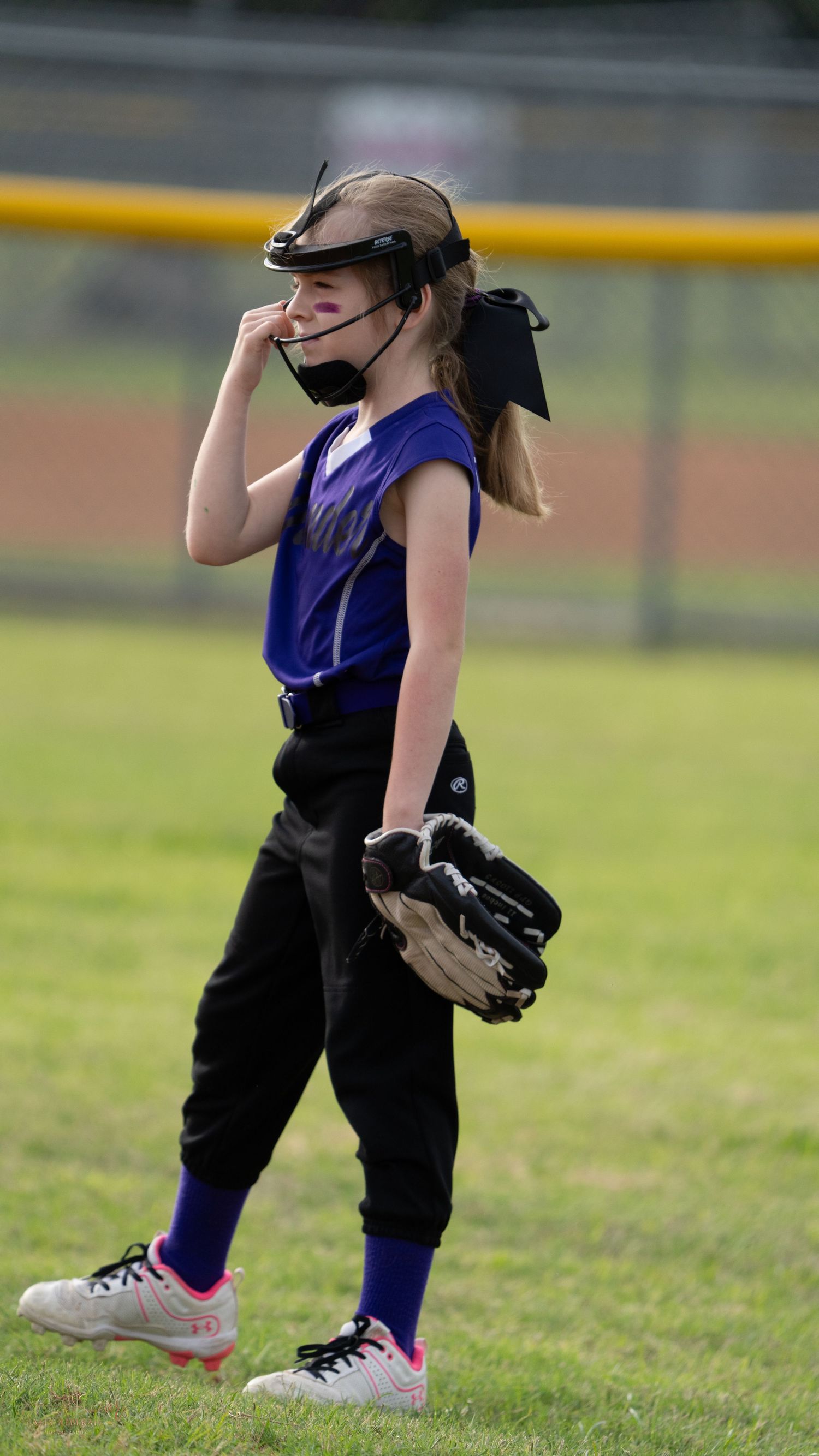 Youth softball player in purple jersey and black pants reacts during an outdoor game on a sunny baseball field.