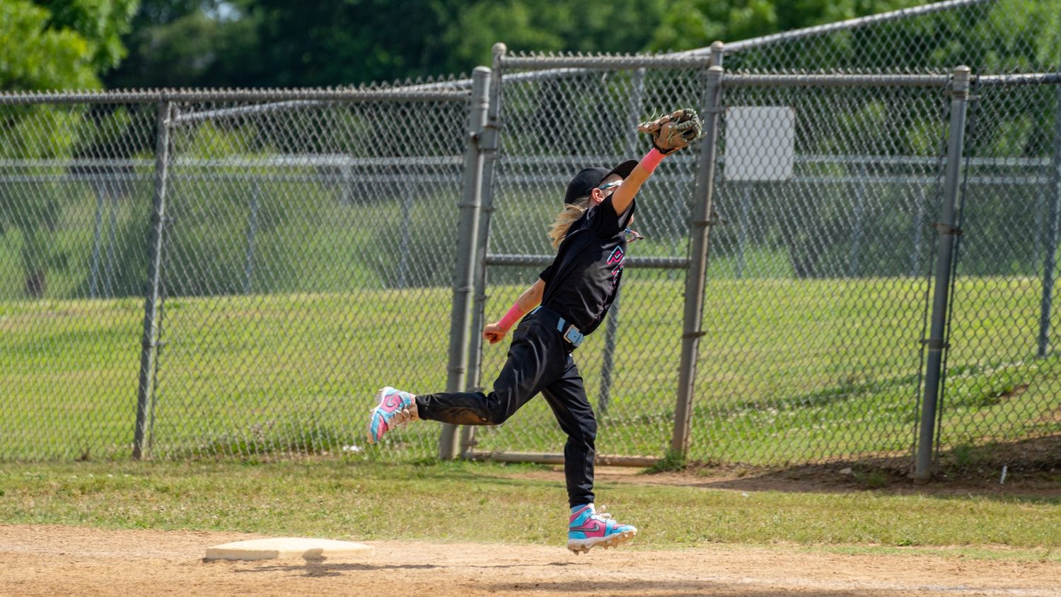 Young baseball player leaping to catch a ball near a chain-link fence on a sunny day.