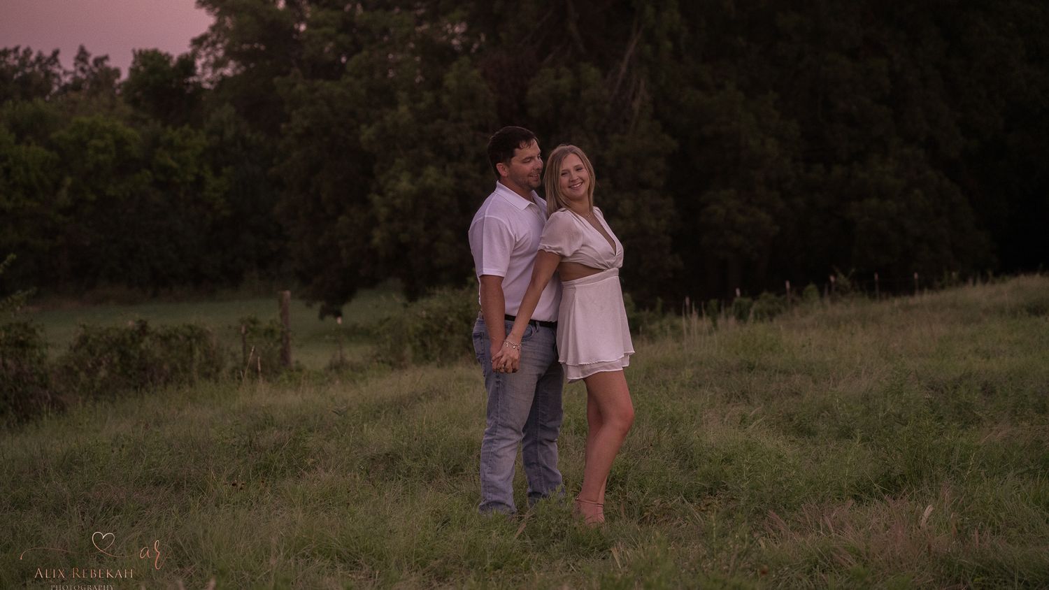A couple shares an intimate moment in a field during dusk lighting.