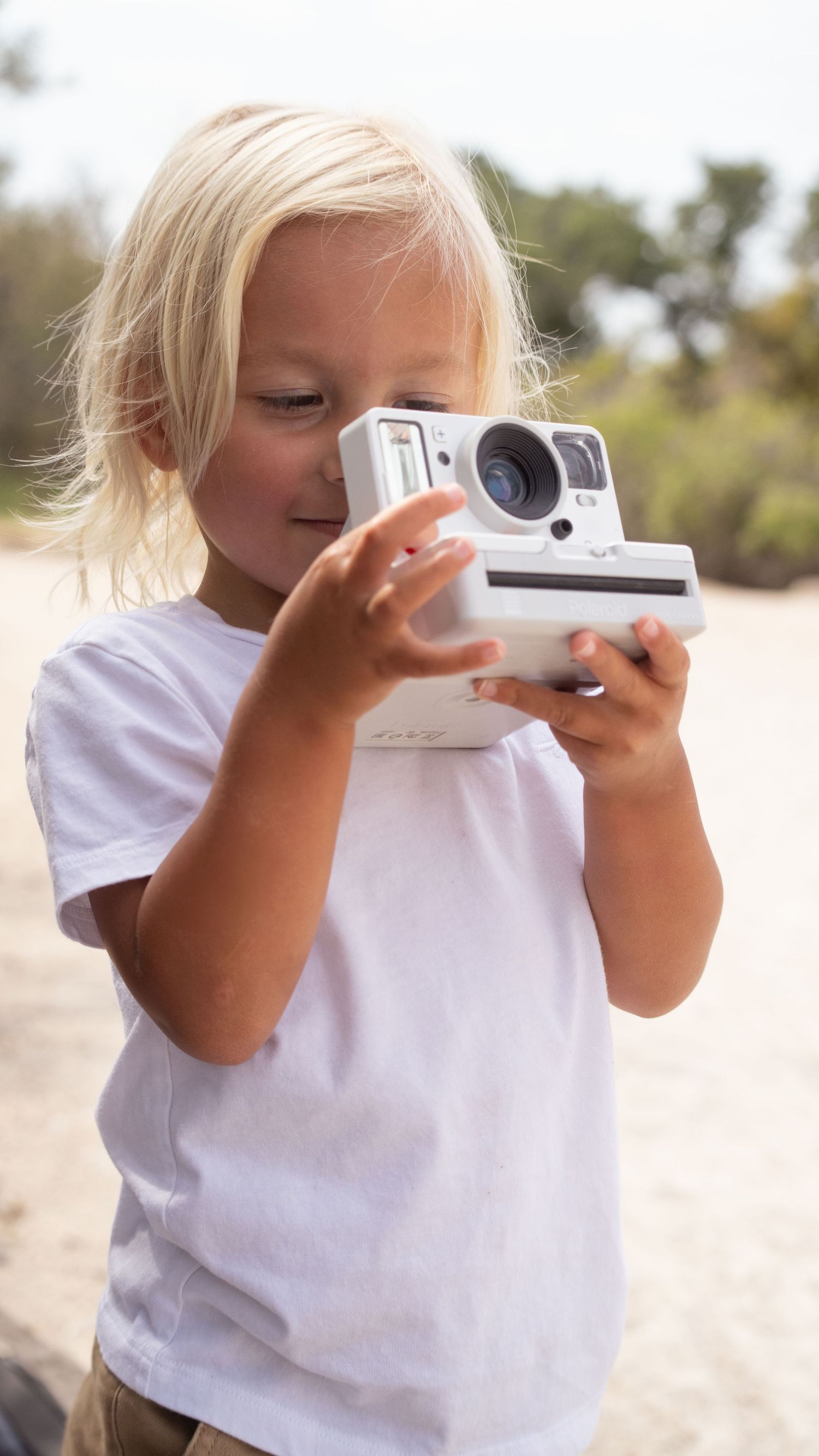 A child in white t-shirt examining an instant camera outdoors.