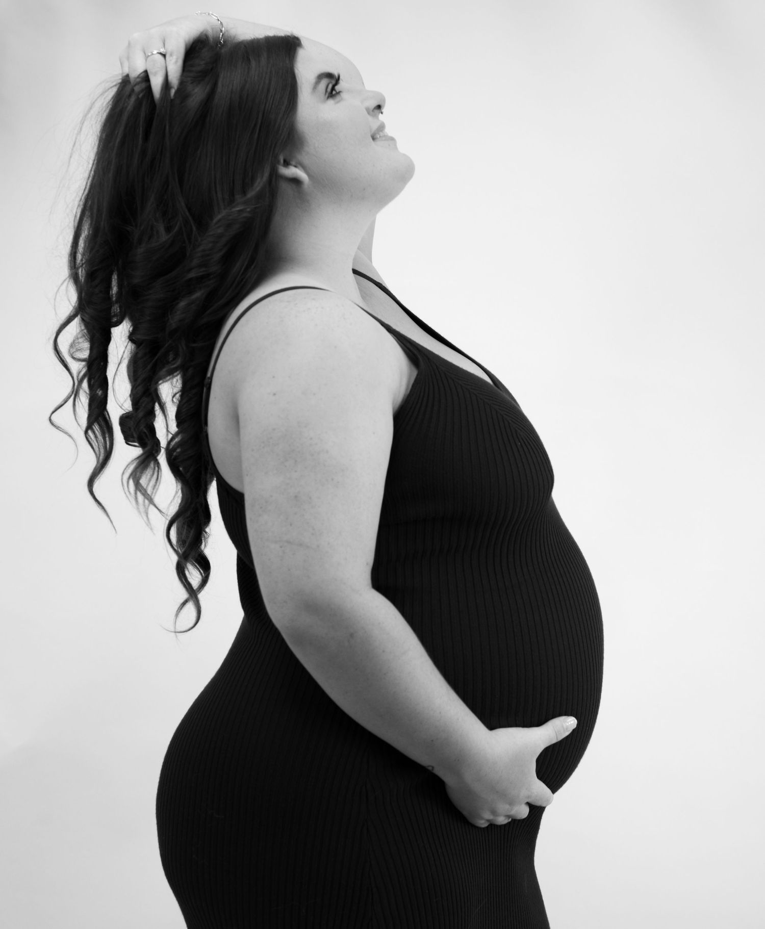 Black and white maternity portrait showing woman in dark top with long curly hair against light background.