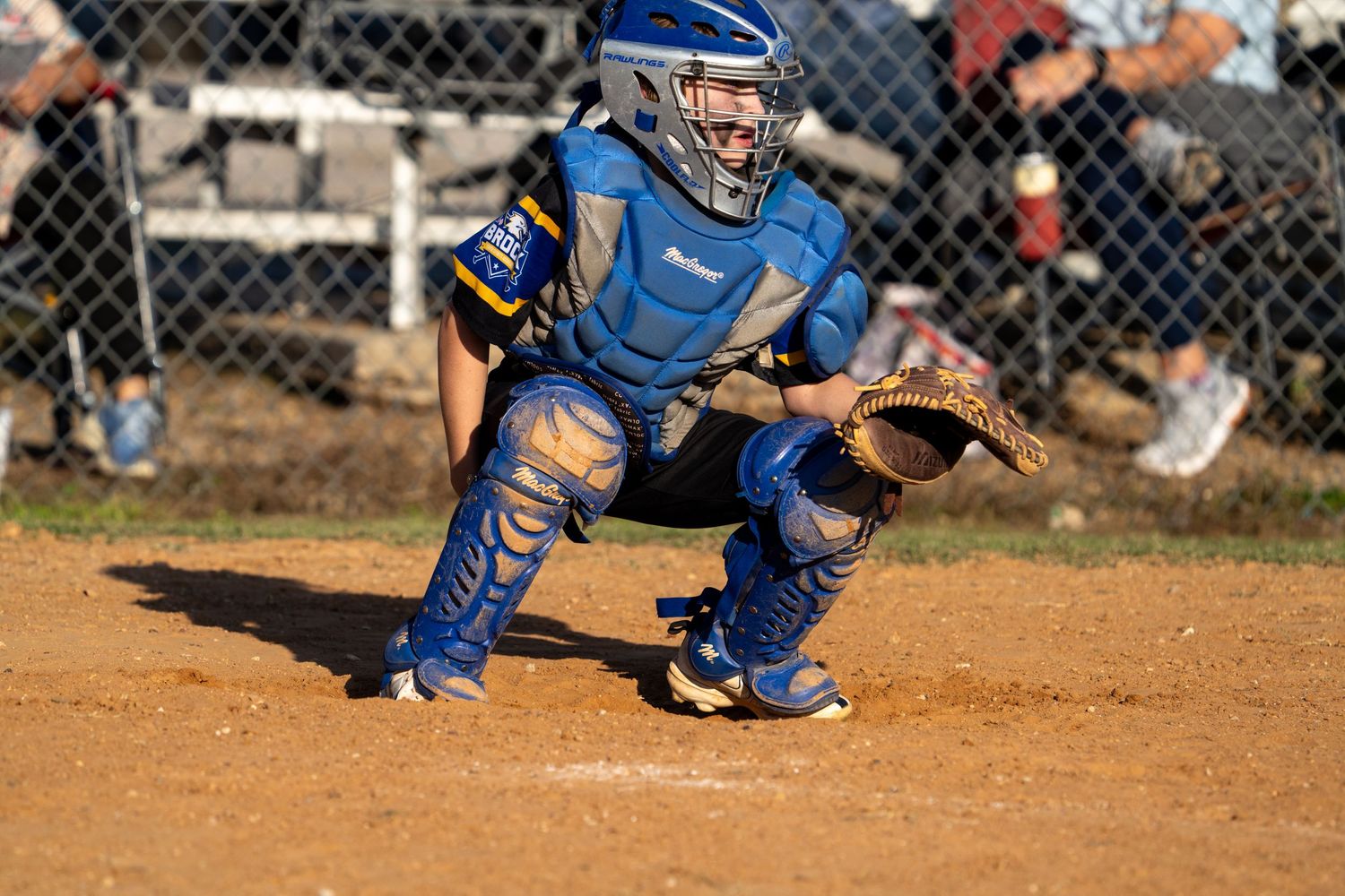 Baseball catcher in blue protective gear crouches in ready position on dusty baseball diamond.