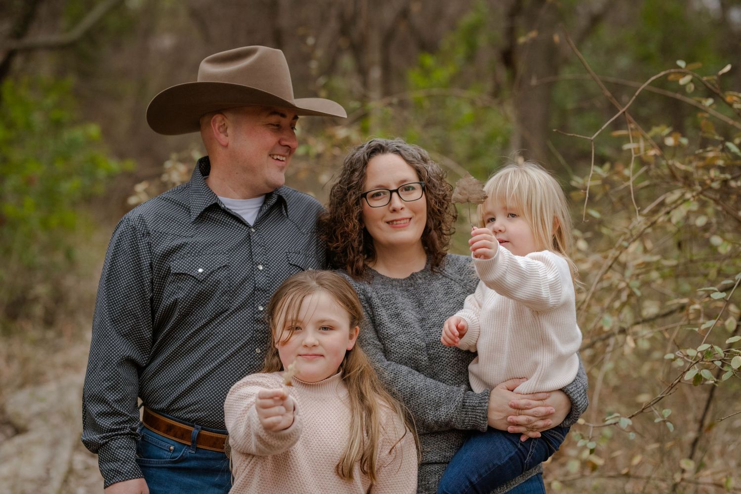 A family of four stands together outdoors amidst autumn foliage in casual attire and rustic setting.