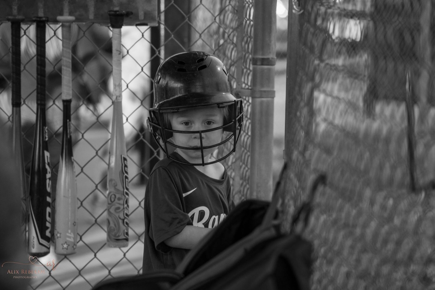 A young baseball player in a batting helmet watches from behind the dugout fence in black and white.