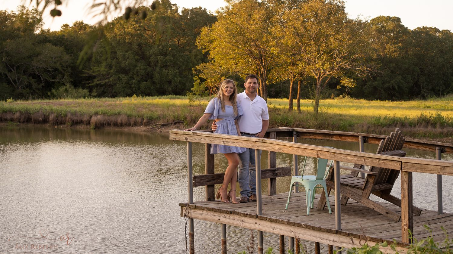 A couple sits together on a wooden dock overlooking a peaceful pond at sunset.