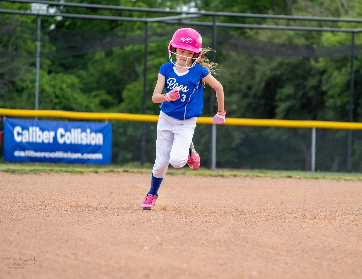 Youth softball player in blue uniform and pink helmet runs between bases on dirt infield during game.