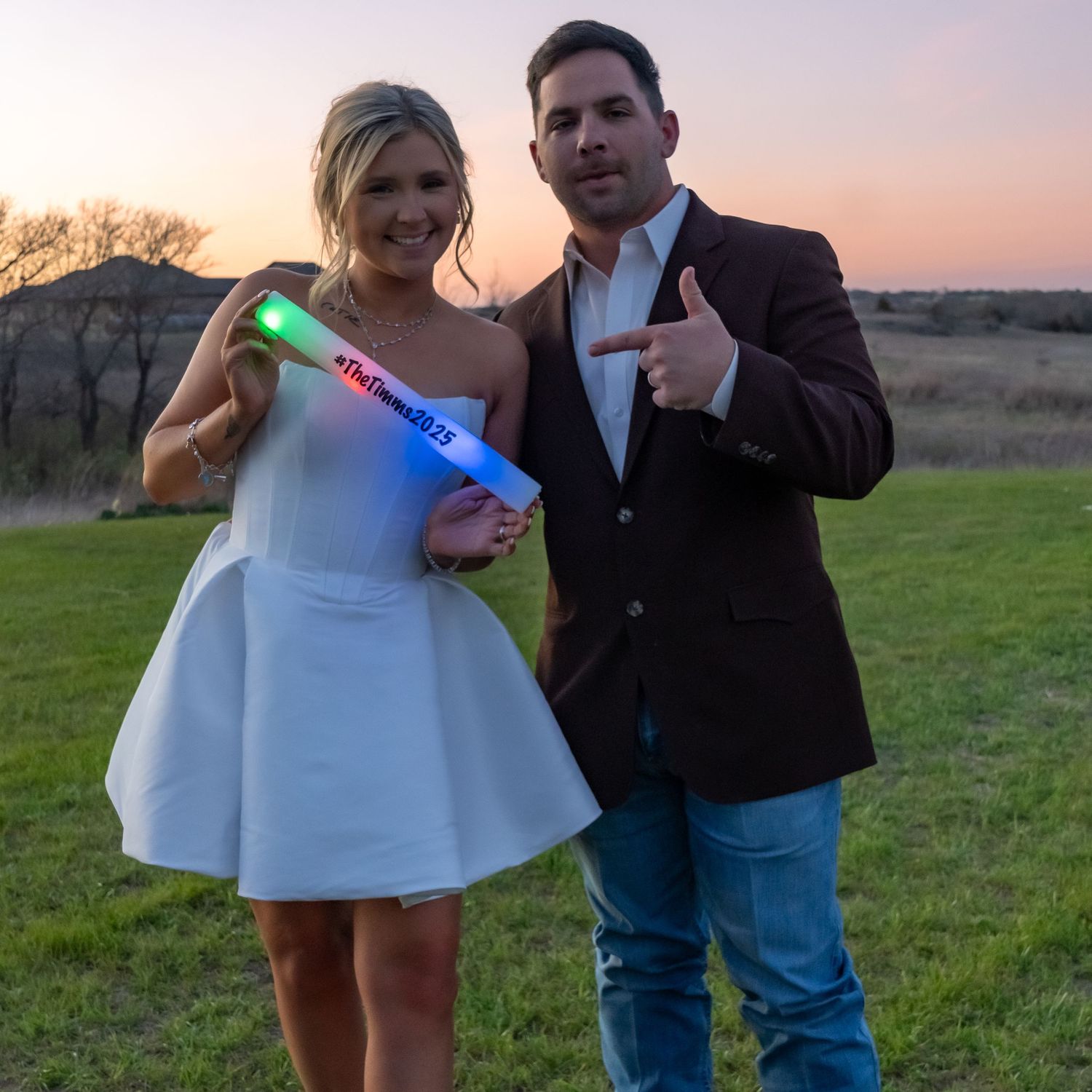 Couple in wedding attire poses at sunset while holding a glowing blue light stick on a grassy lawn.
