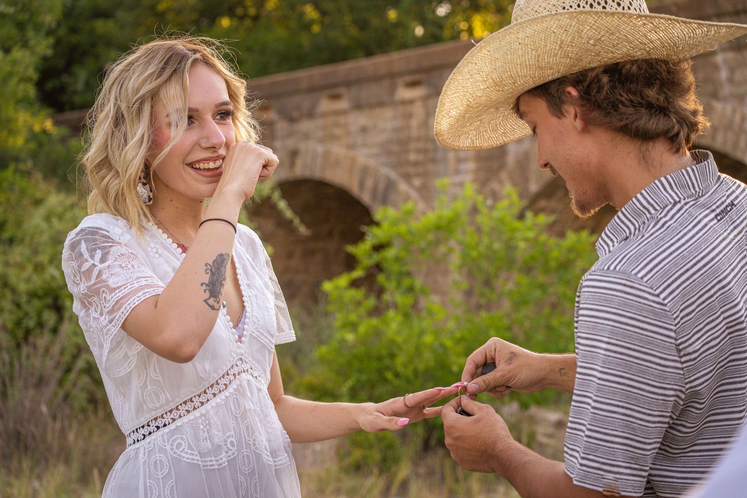 A romantic outdoor proposal scene with a person in a white lace dress receiving a ring from someone wearing a cowboy hat.
