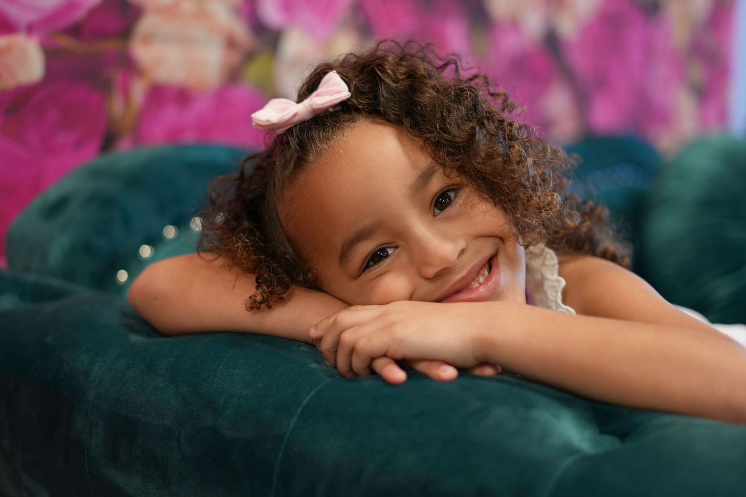 Relaxed pose on teal velvet couch with pink floral backdrop and natural curls.