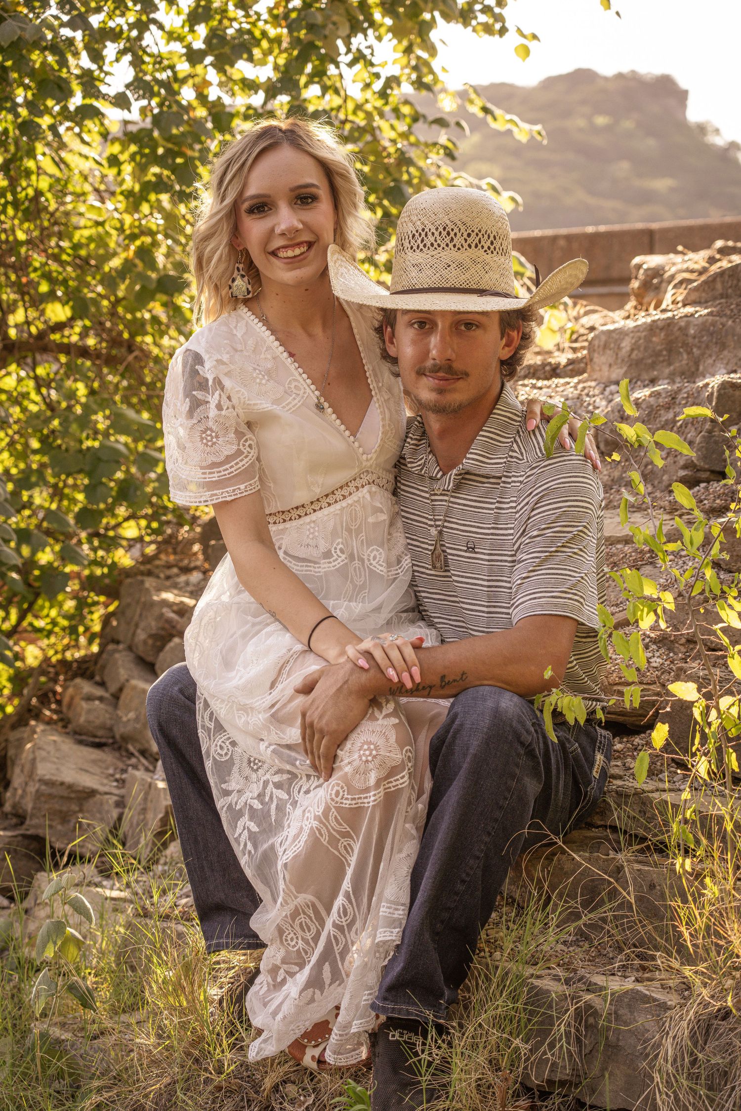A couple in casual country attire sits close together on rocky terrain during golden hour sunset.