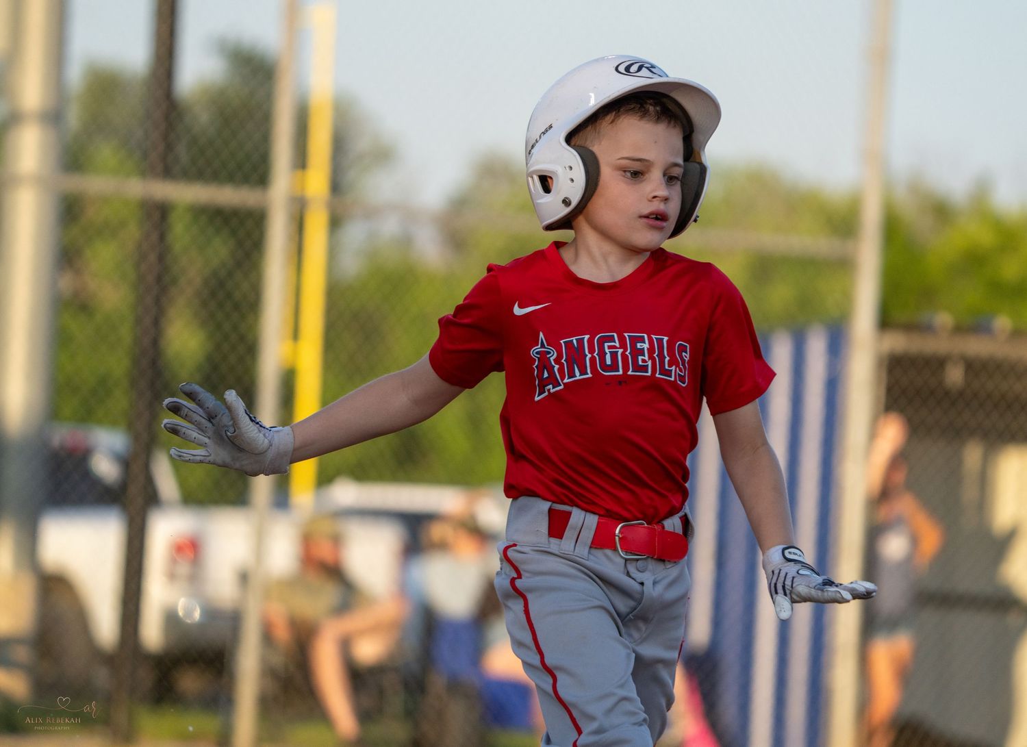 Youth baseball player in grey uniform swings bat during little league game with teammates and spectators looking on.