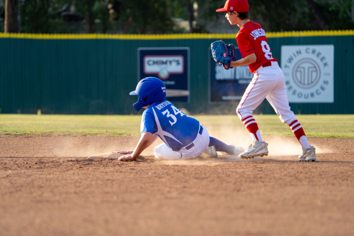 Baseball player in blue slides into base as fielder in red attempts to make the play during youth game.