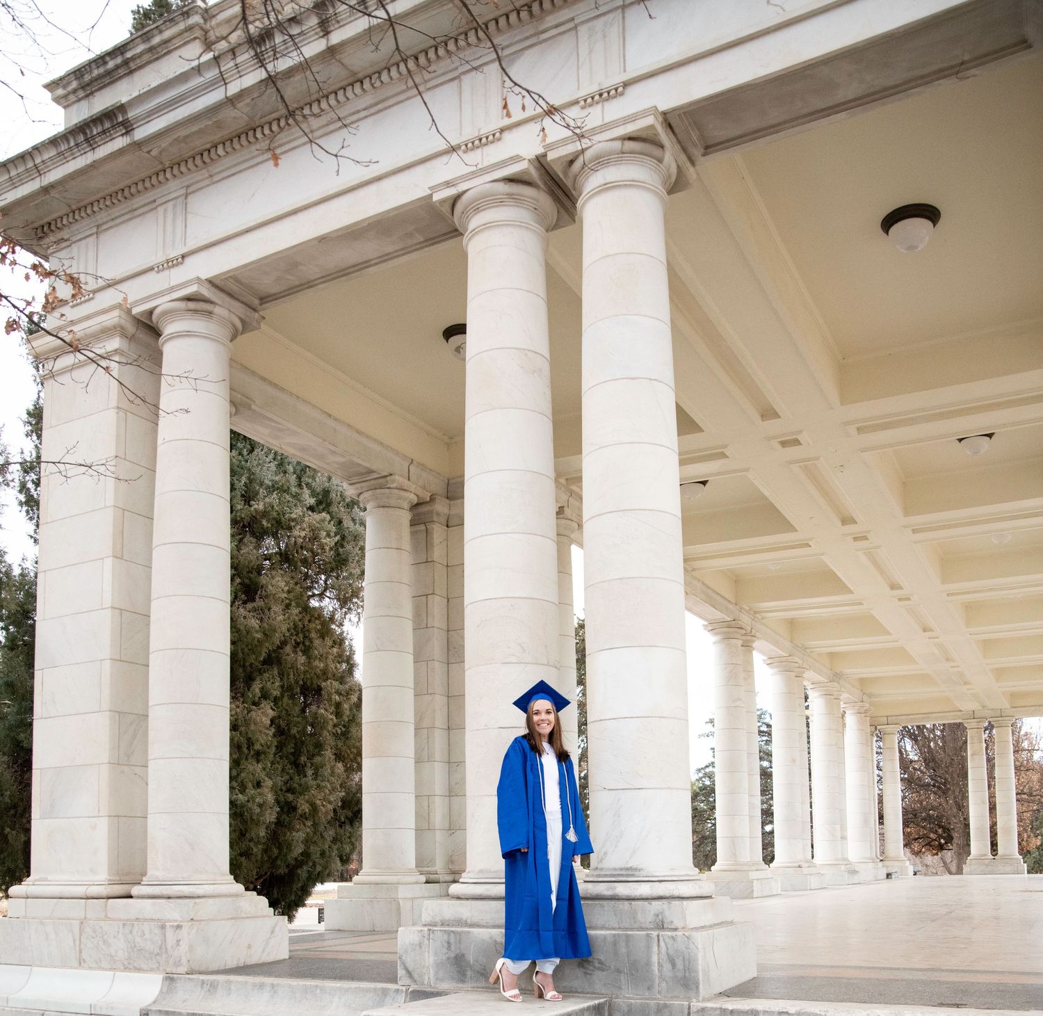 Graduate in blue cap and gown poses between classical white columns.