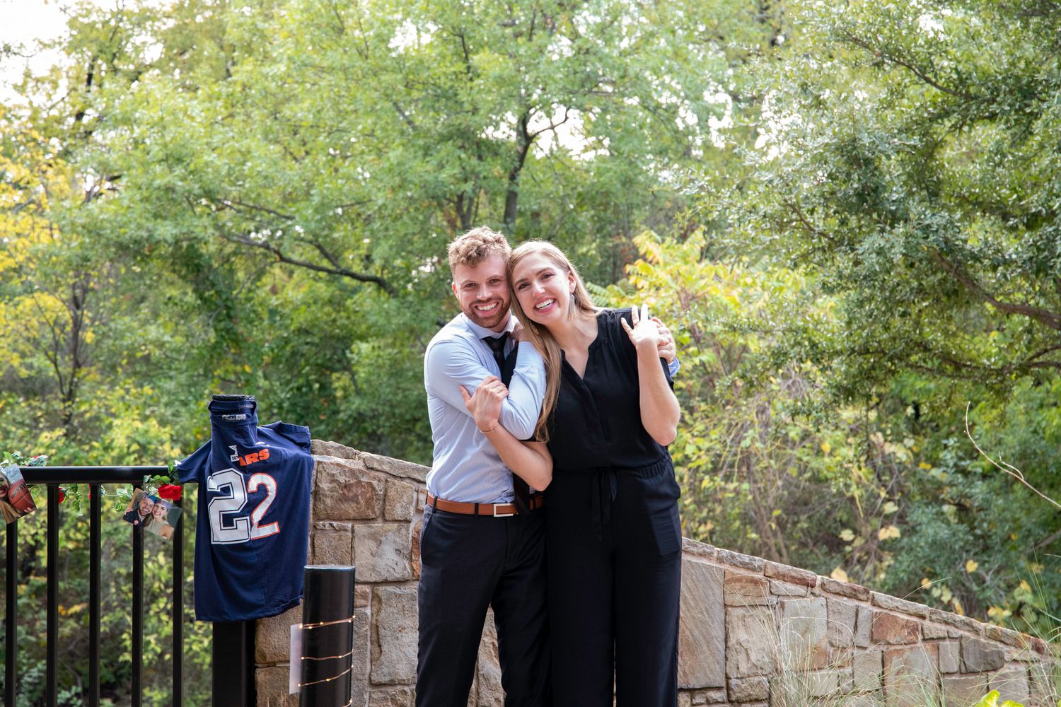 A couple poses together on a stone patio with trees in the background and a sports jersey hanging nearby.