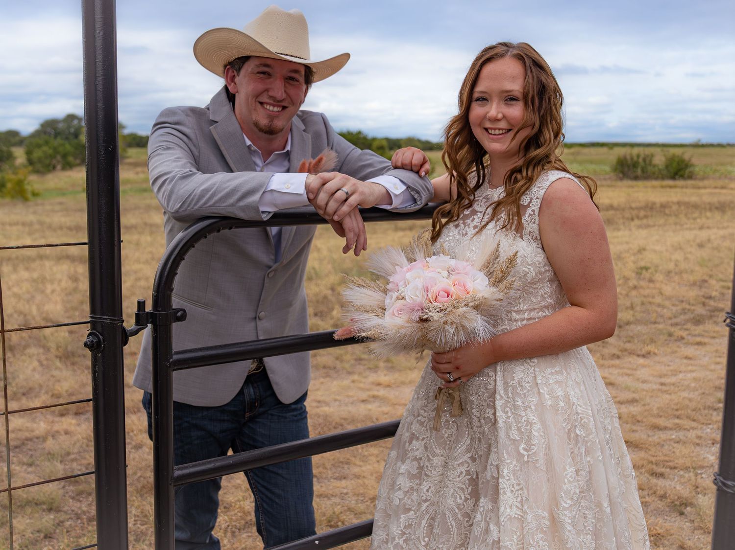 Bride in lace gown holding pink bouquet poses with groom in cowboy hat at ranch gate in open field.