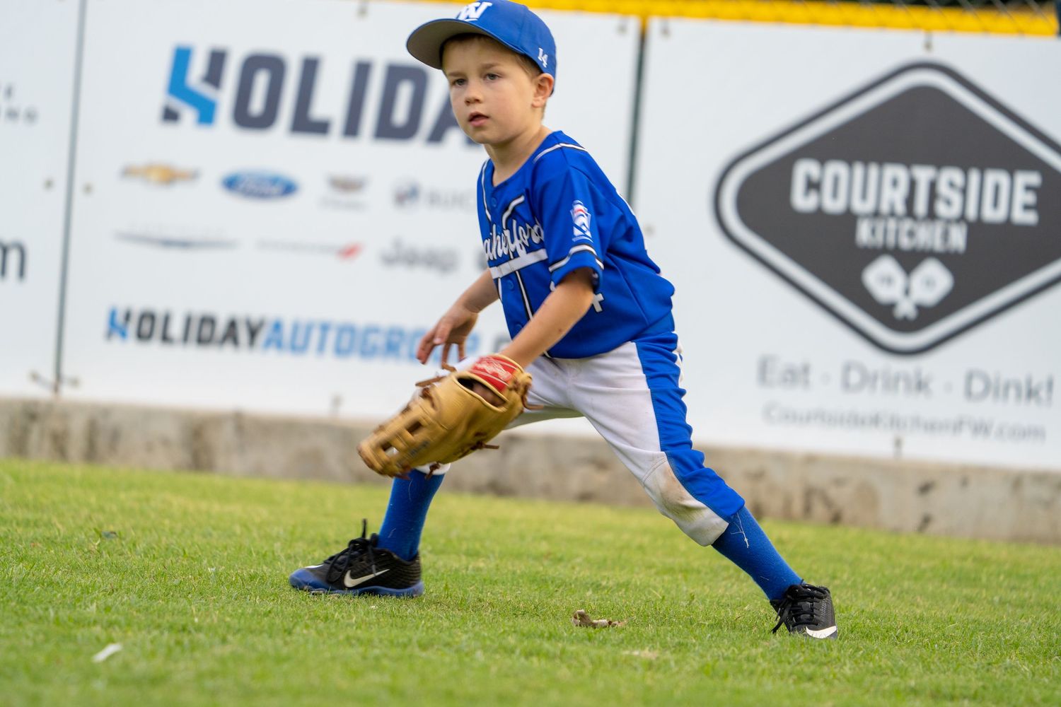 Young baseball player in blue uniform crouches in fielding position on grass field with advertising banners in background.