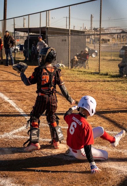 Youth baseball catcher in orange gear calls a play while runner in red jersey slides into home plate on dirt field.