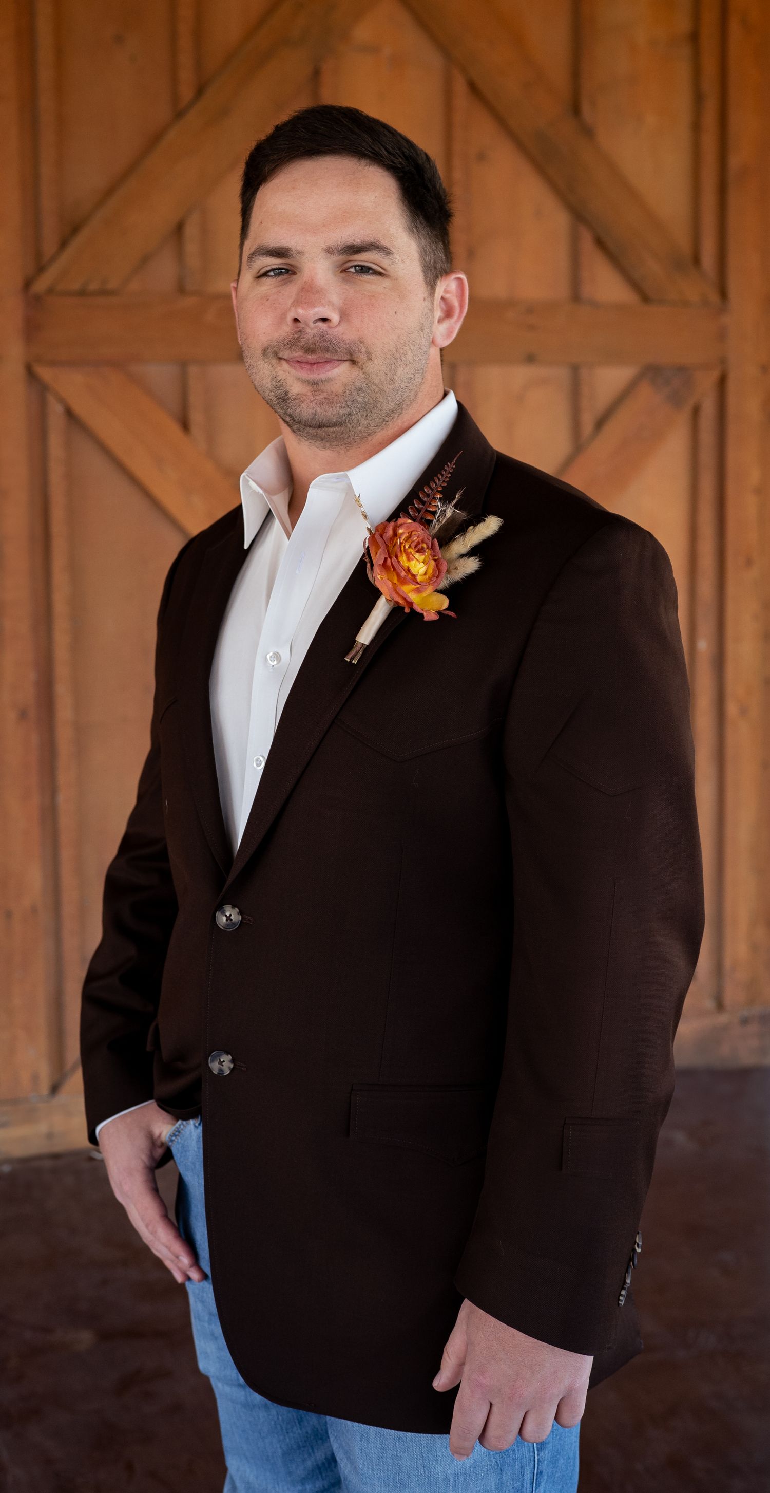 A groom in a dark brown blazer with orange boutonniere stands in front of rustic wooden doors in a casual formal style.