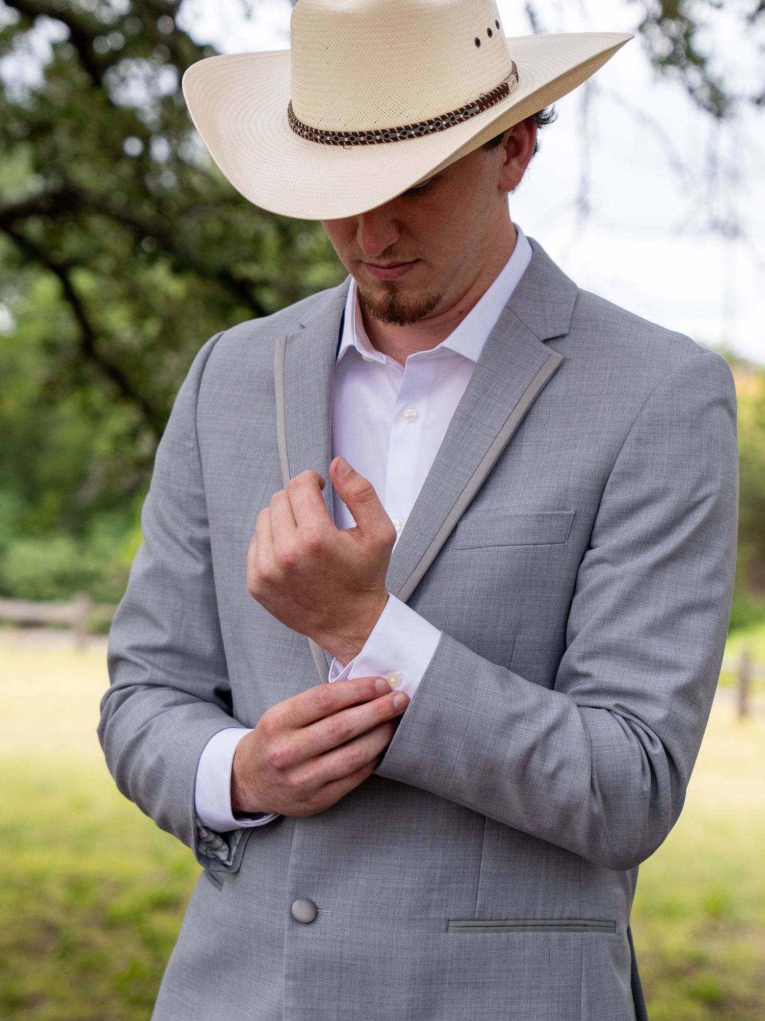 Man in gray suit and cowboy hat adjusting cufflinks outdoors at a rustic ranch setting.