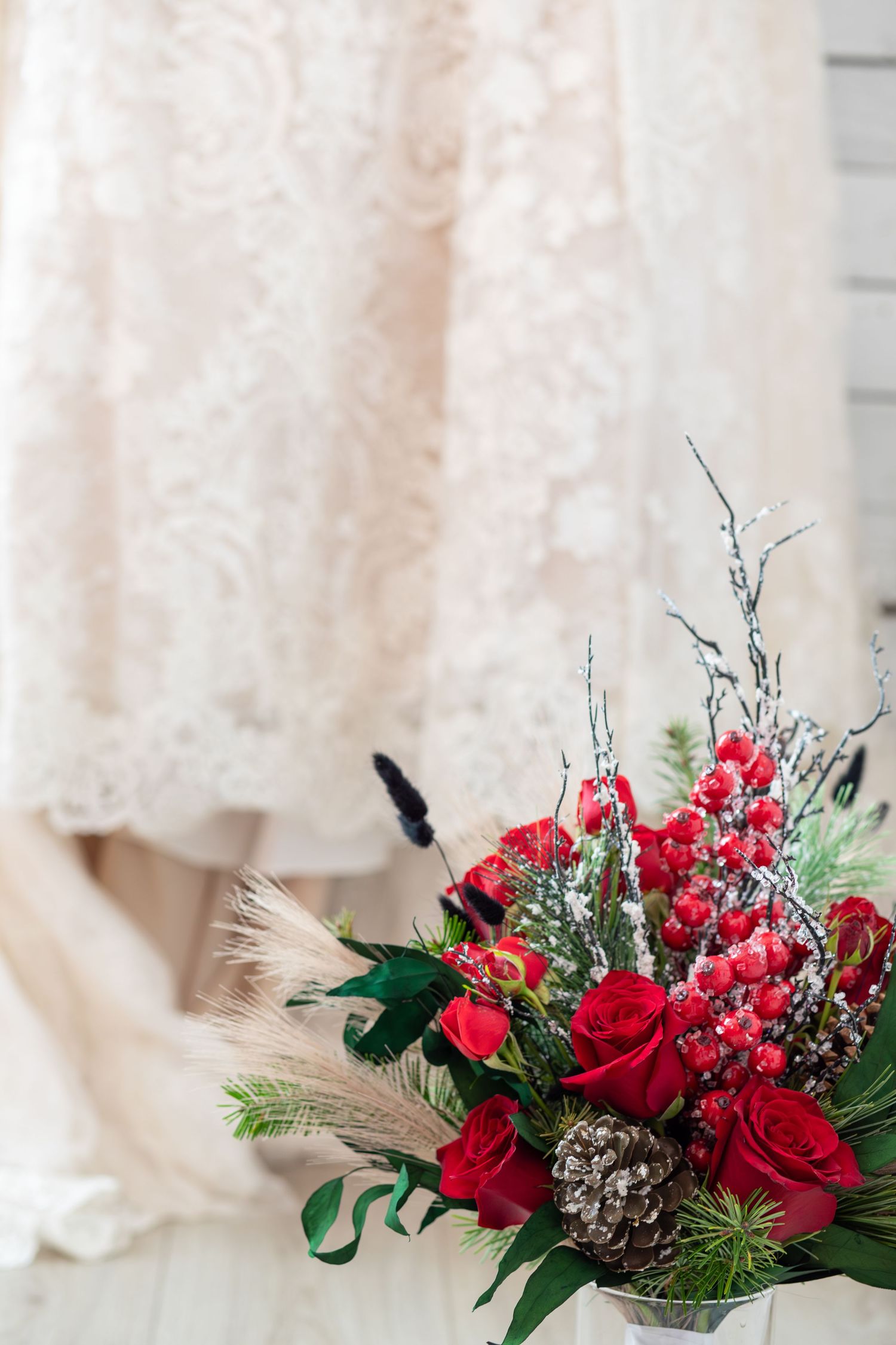 Winter wedding bouquet with red roses, berries, pine cones, and greenery in front of a lace bridal gown.