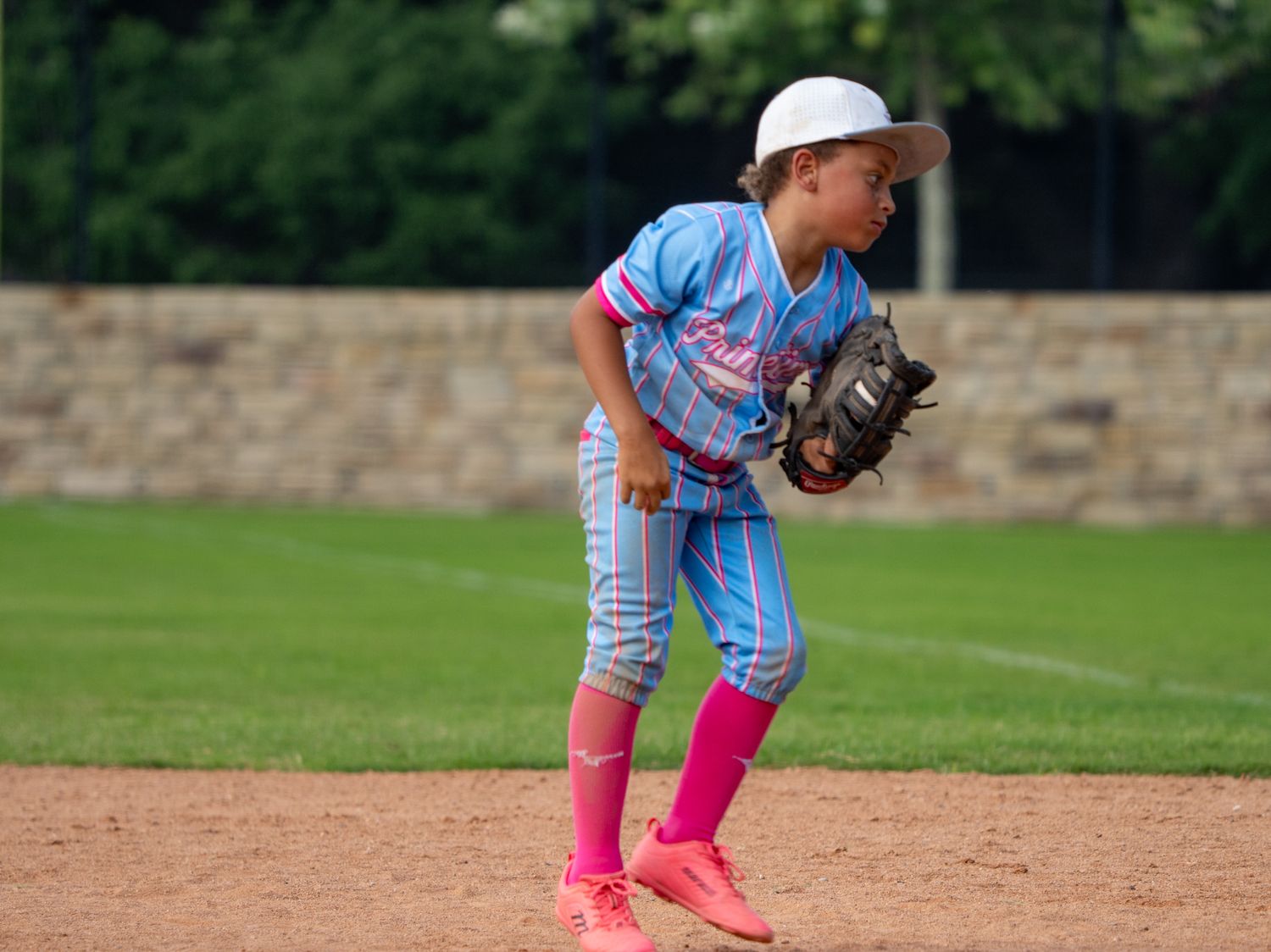 Young baseball player in blue and pink uniform fielding position on dirt infield during little league game.