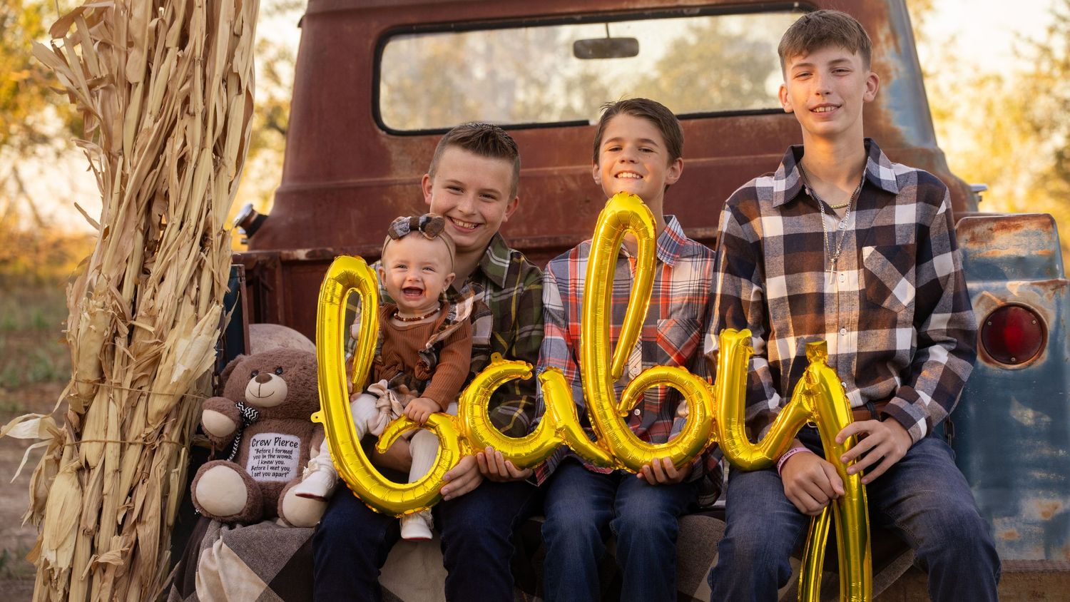 Family sits on vintage red truck with gold balloon letters spelling 'baby' during autumn photo session.