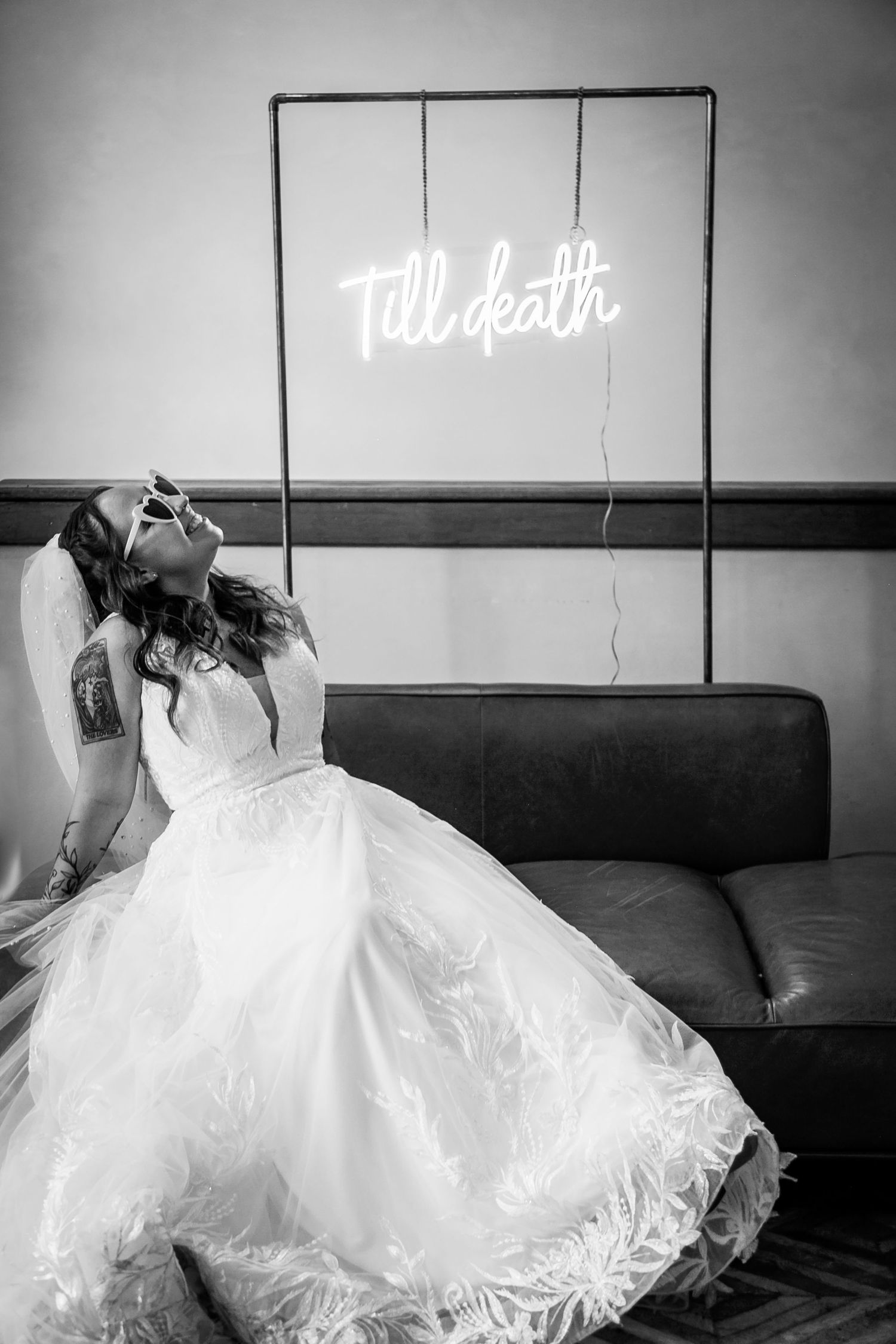 Bride in white dress reclines beneath neon 'Till death' sign in black and white photography.