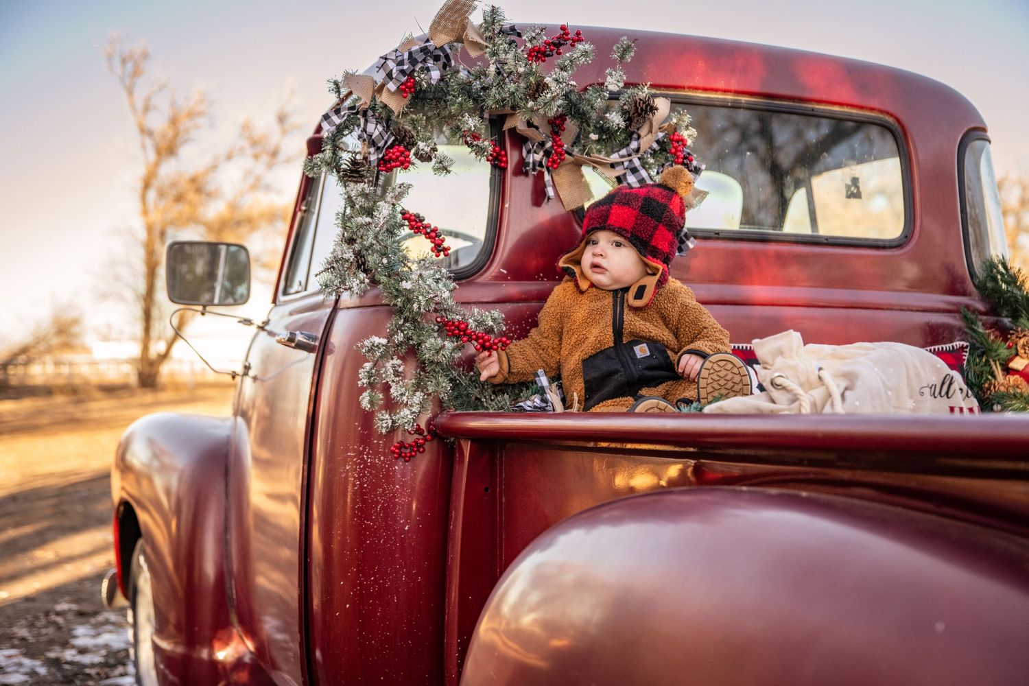Vintage red truck decorated with Christmas garland and baby in cozy brown winter outfit.
