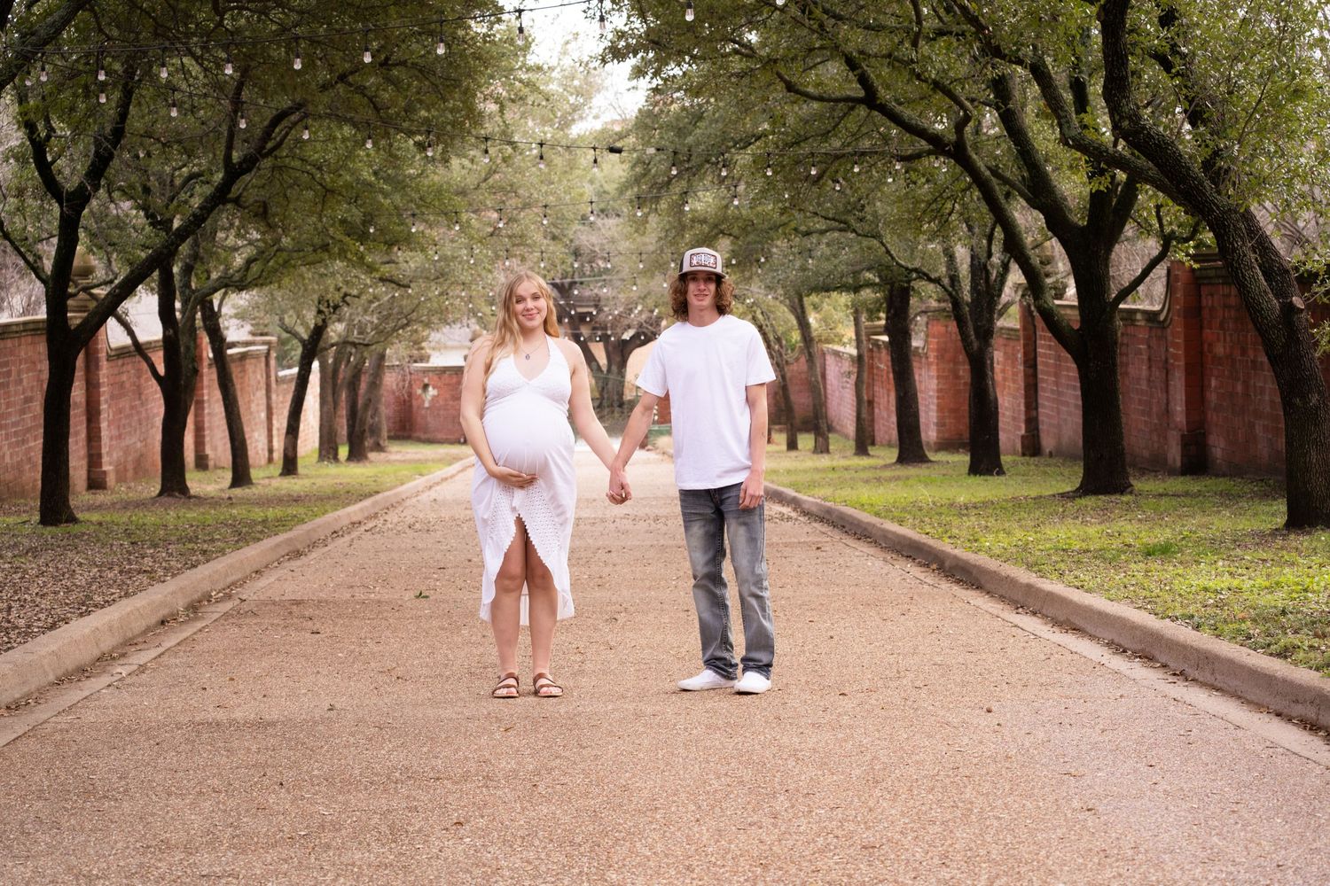 Expectant couple walking hand in hand down a tree-lined path.