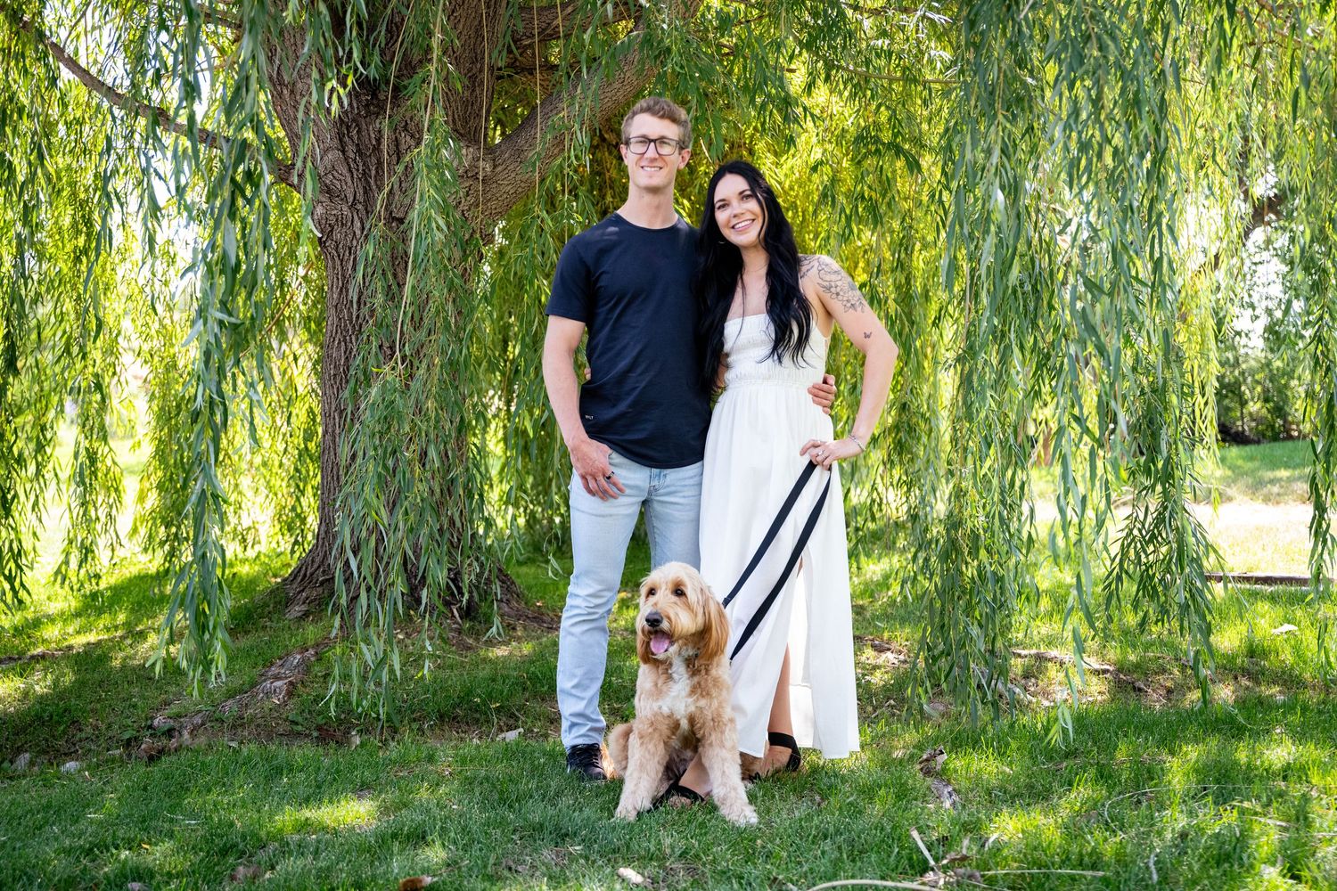 Happy couple poses with their goldendoodle dog under willow trees in park setting.