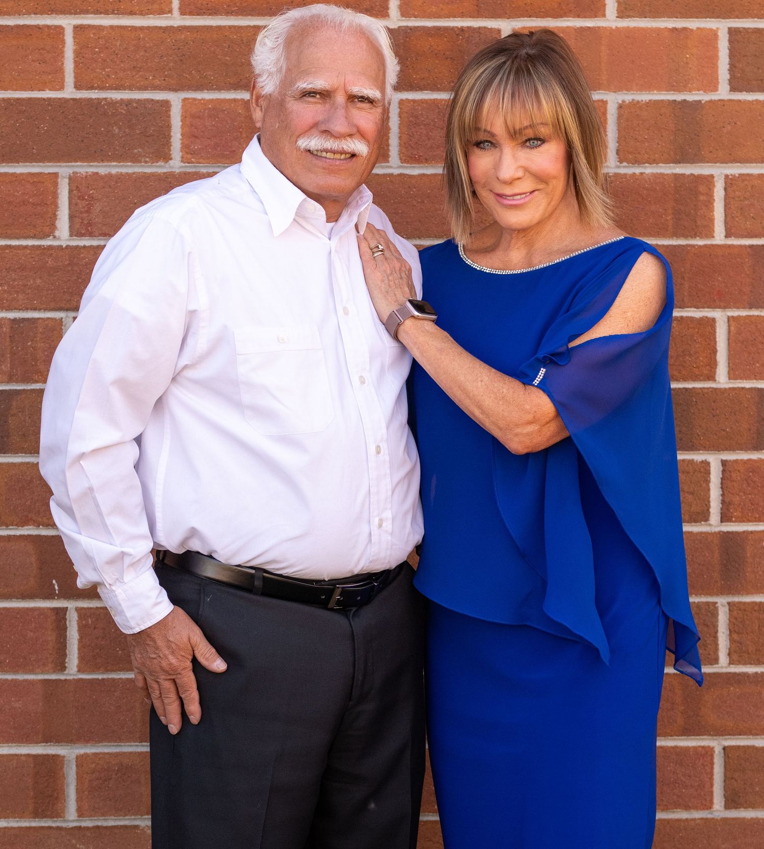Couple poses against brick wall wearing a white dress shirt and royal blue dress.