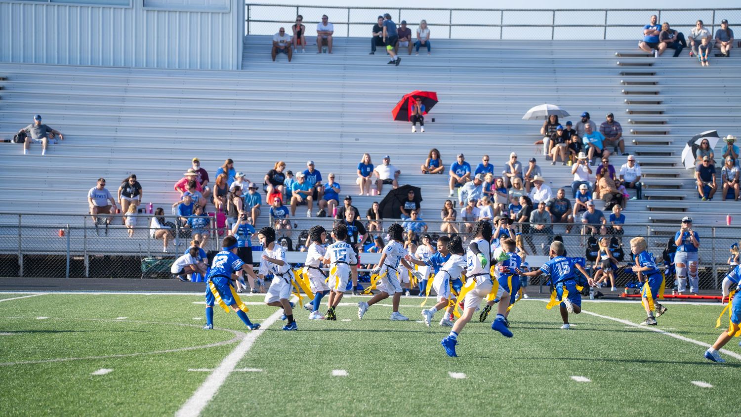 Youth football teams compete in a game on a field with stadium bleachers in the background.