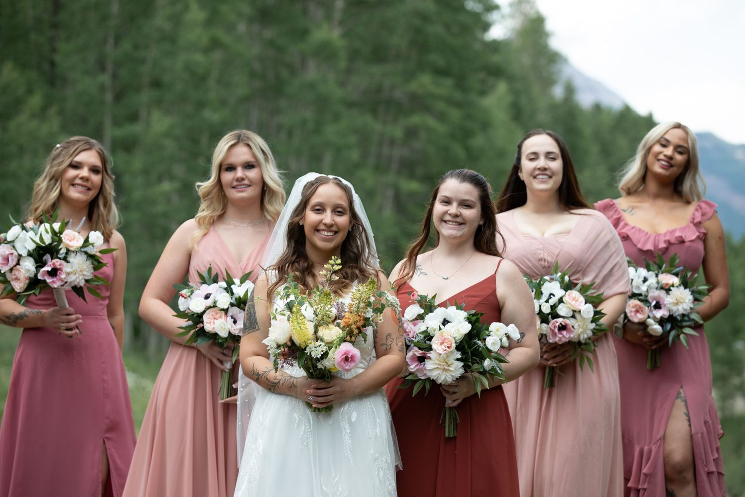 Bridesmaids in dusty rose dresses pose with bouquets against forest backdrop.