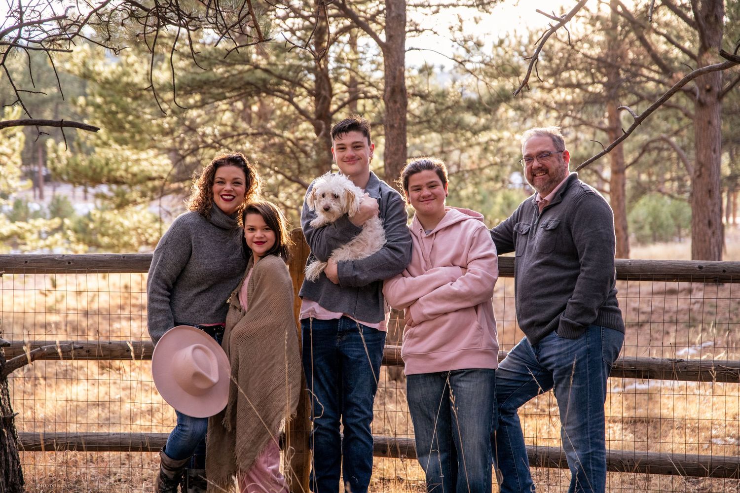 A group gathered in a pine forest with a small white dog and rustic fence in the background.