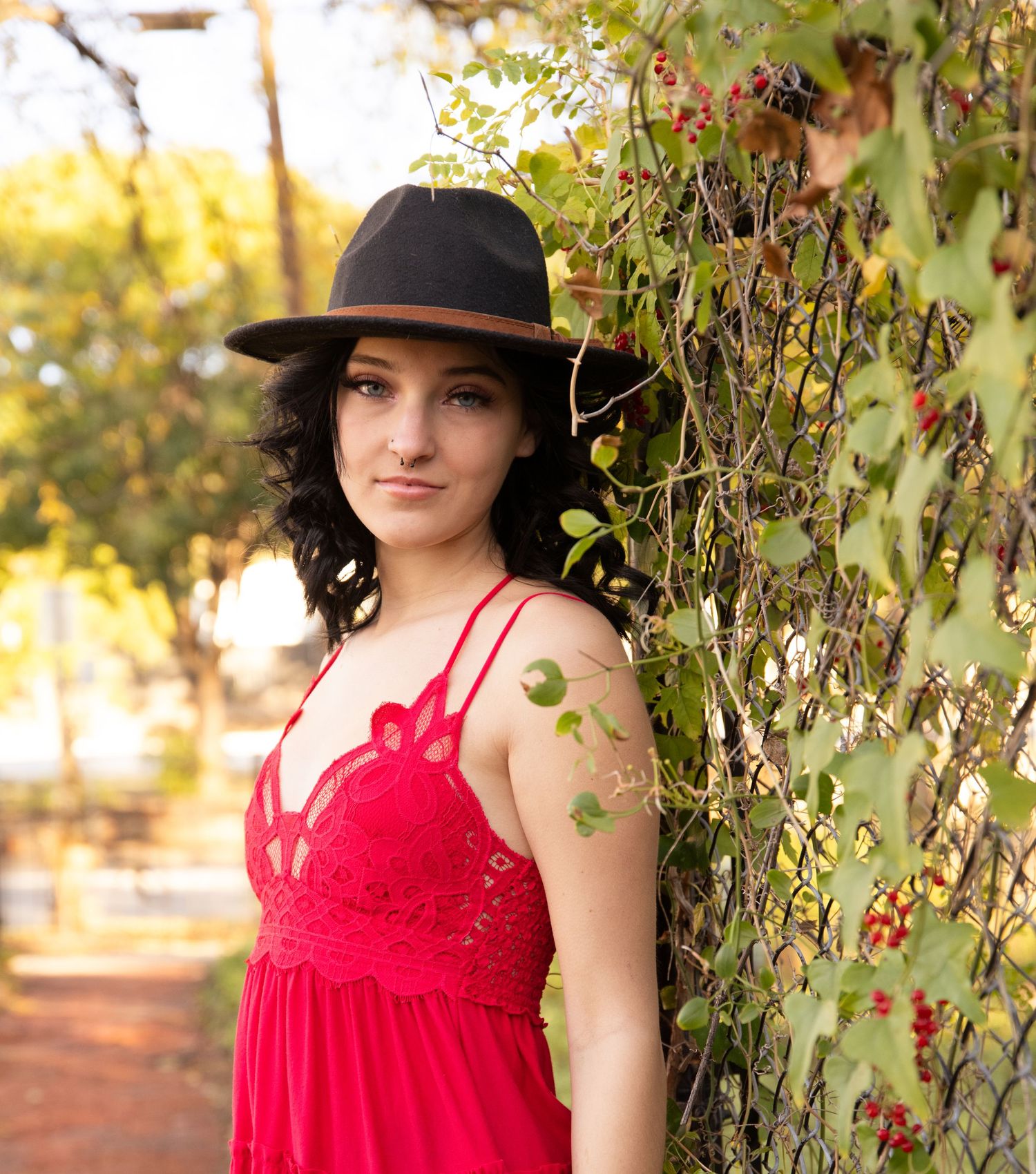 Woman in red dress and black hat poses against leafy backdrop in autumn light.