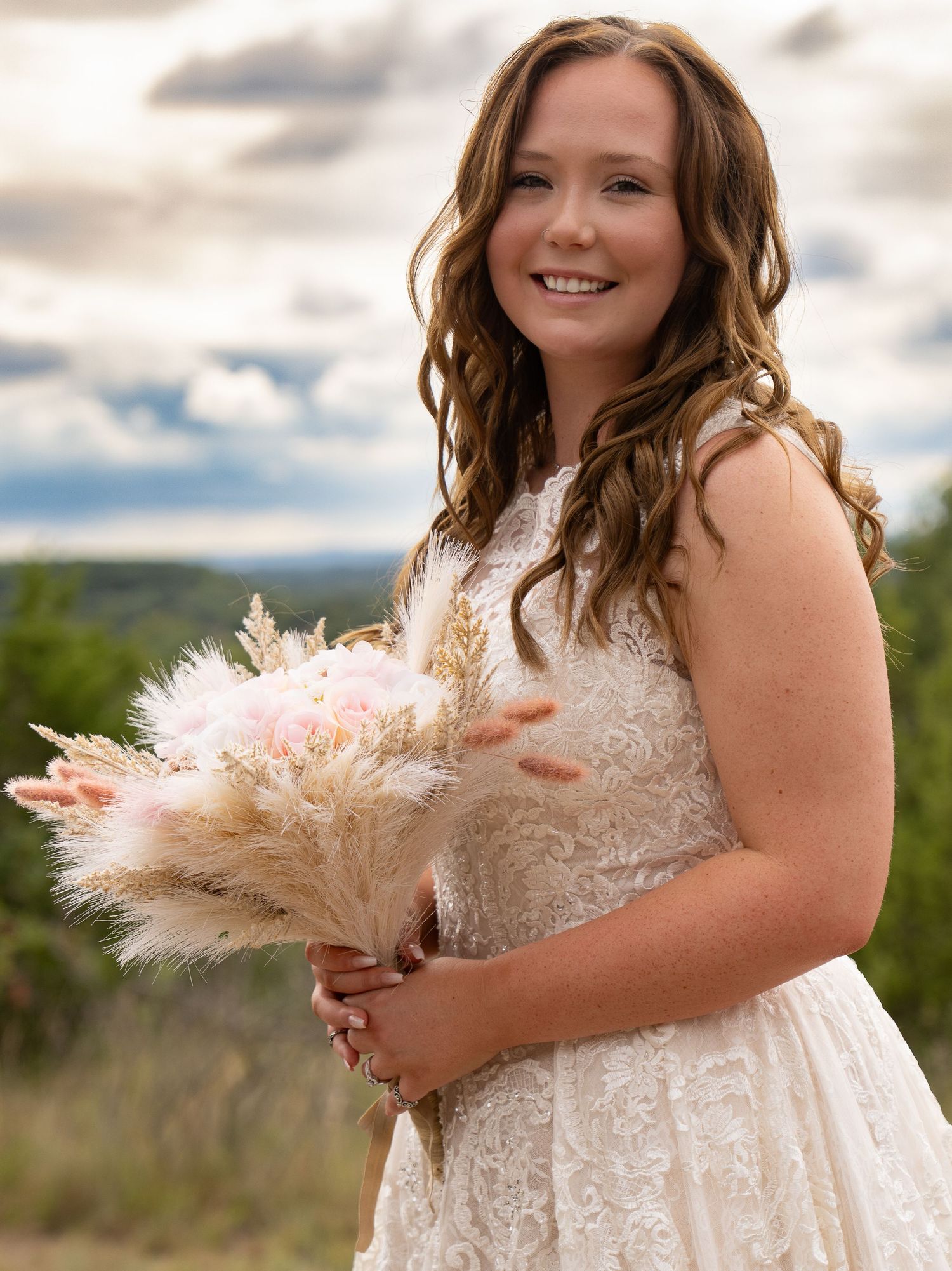 Young bride in lace dress holds pampas grass bouquet outdoors under cloudy sky.