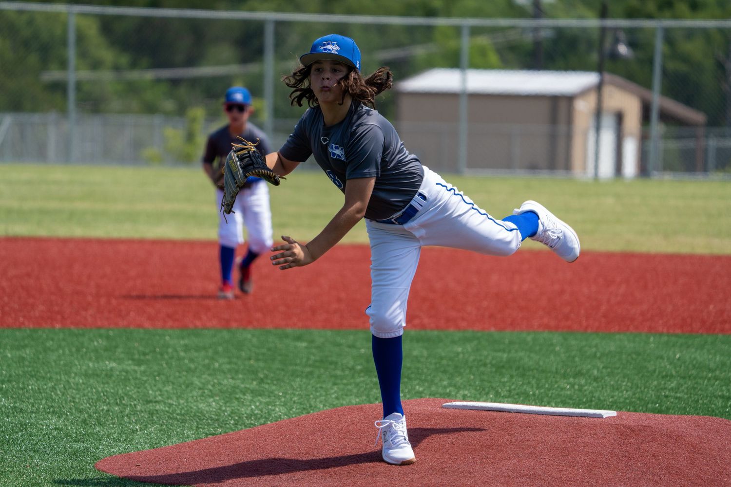 A youth baseball pitcher follows through their delivery on a bright red artificial turf pitching mound during a game.
