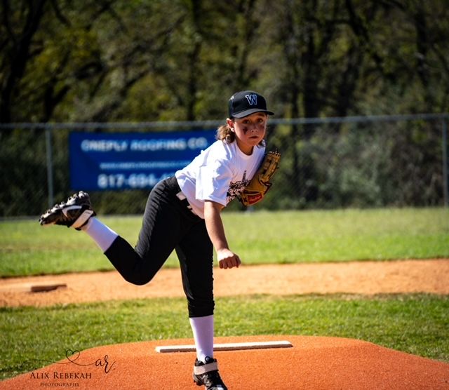 Young baseball pitcher in white and black uniform follows through on delivery from pitcher's mound during game.