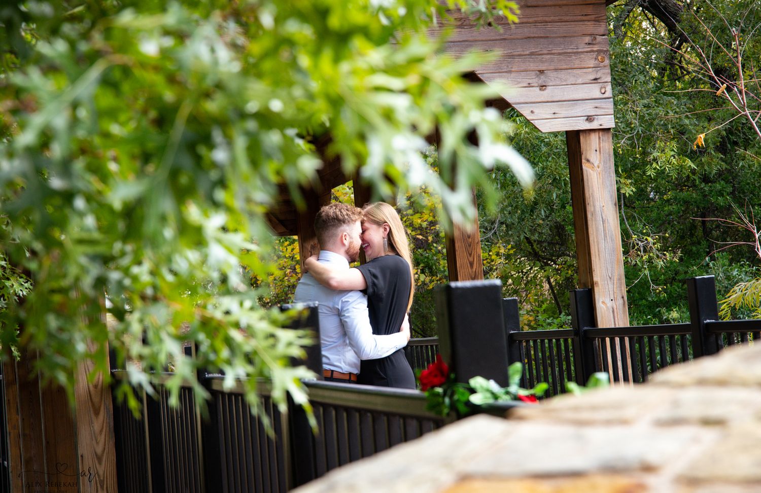 A white railing extends along a wooden deck surrounded by lush green foliage in a backyard setting.